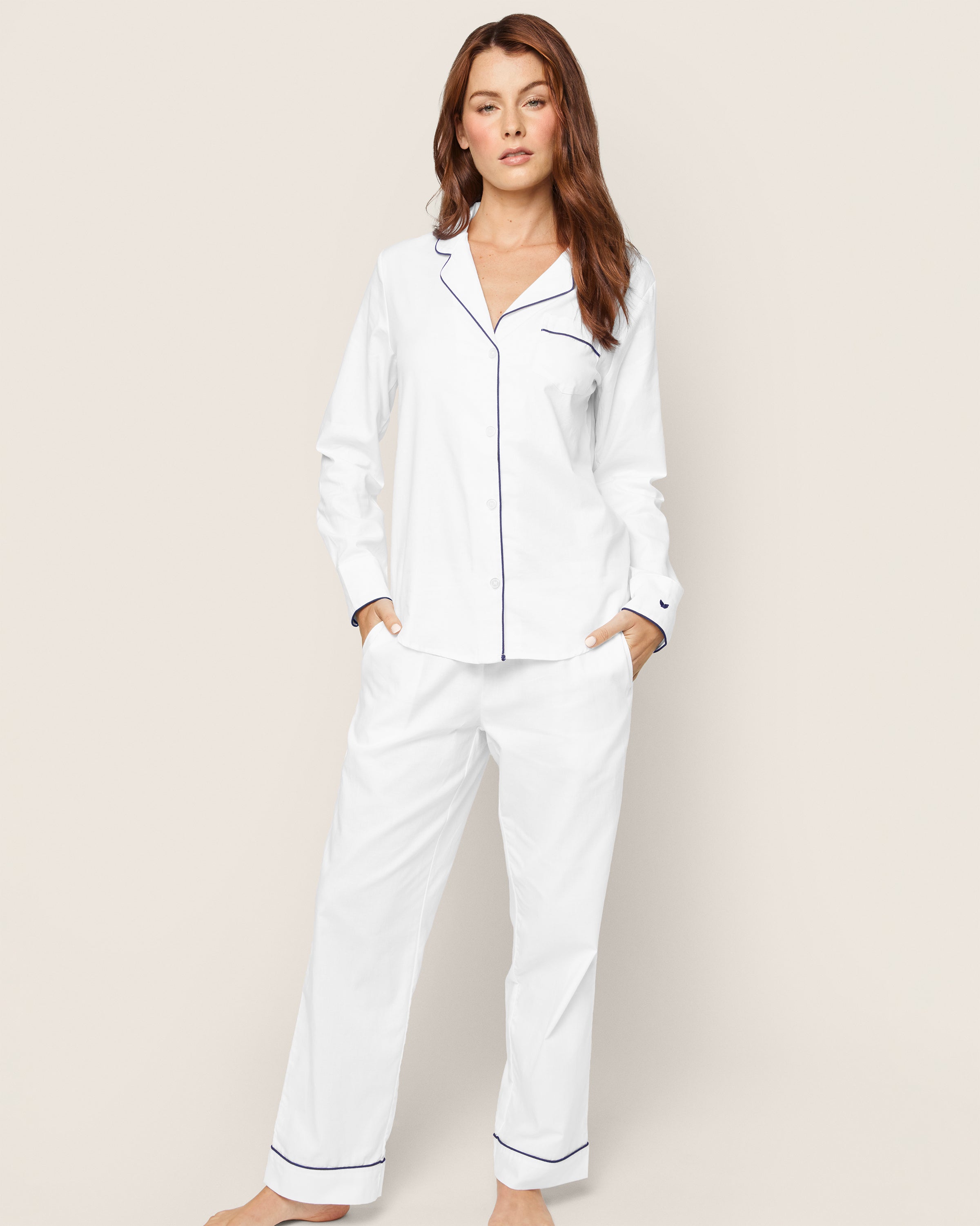 A person in Petite Plumes Womens Twill Pajama Set in white with navy piping stands against a plain background. The top has a collar and buttons, and the loose-fitting, moisture-wicking pants have pockets. They hold a neutral expression with one hand in their pocket.
