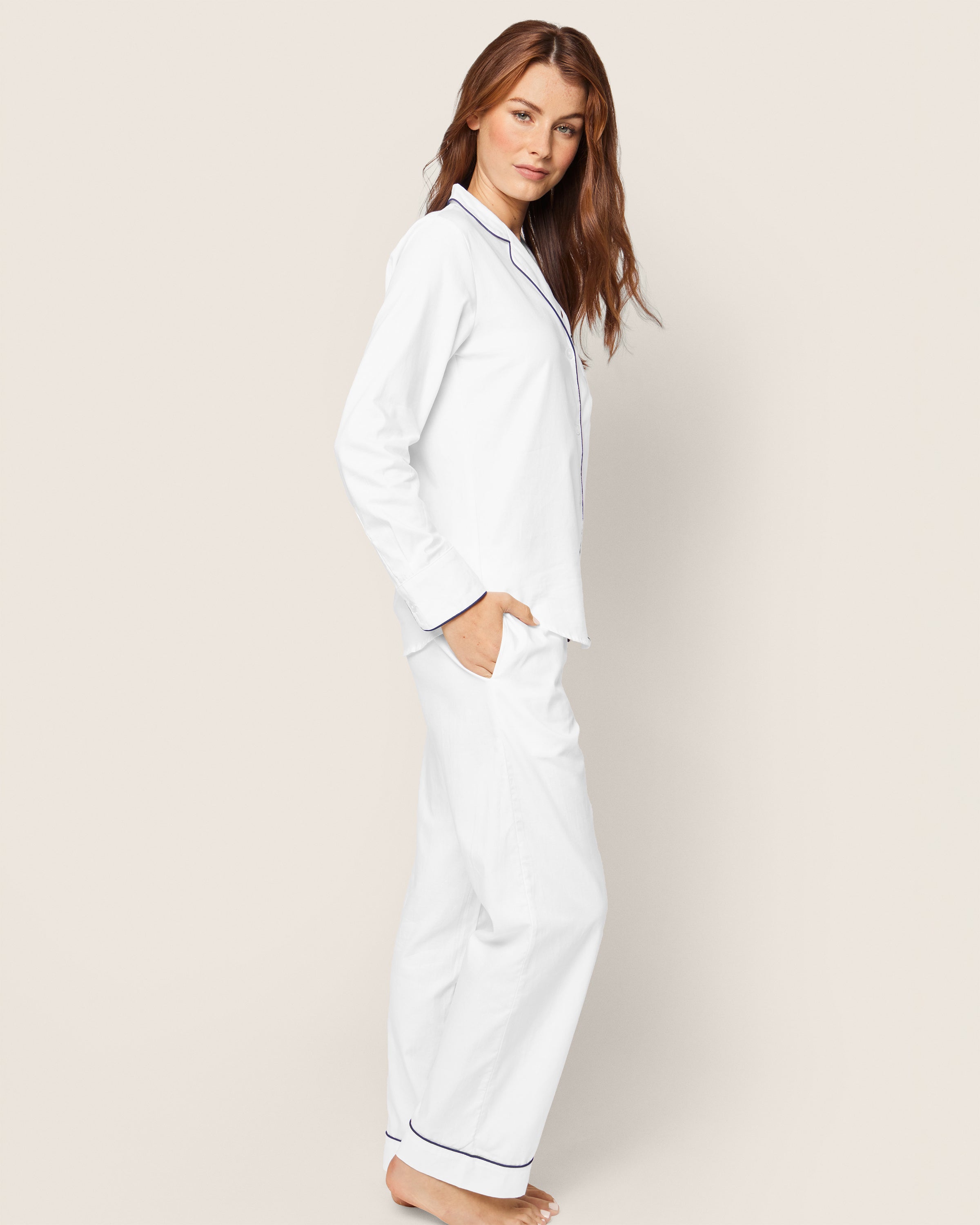 Barefoot with long hair, a person faces forward against a light beige backdrop, casually tucking one hand into a pocket while wearing Petite Plumes Womens Twill Pajama Set in White with Navy Piping.