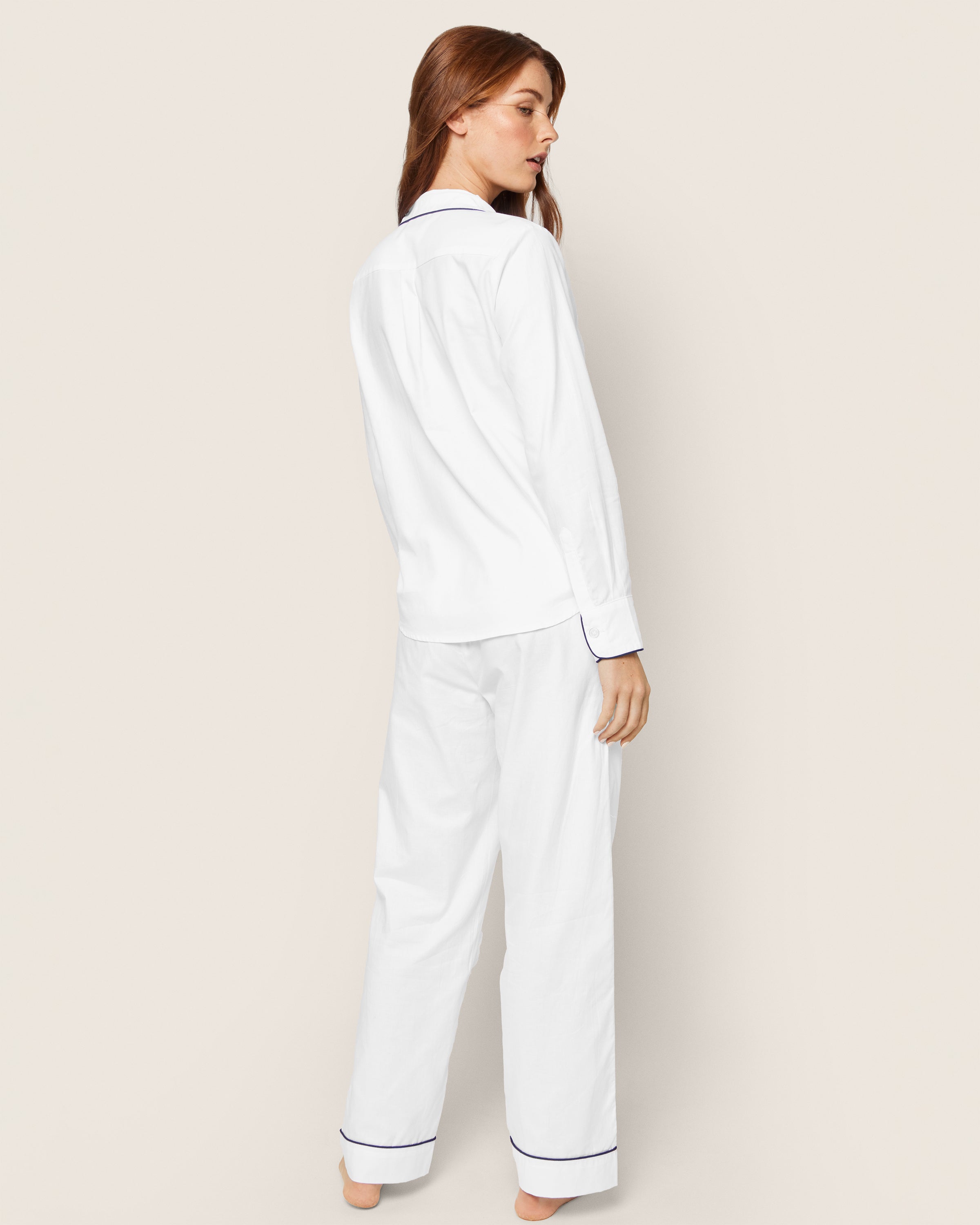 A person with long hair stands facing away in a Womens Twill Pajama Set by Petite Plume. This white set with navy piping features a collared, long-sleeve top and loose pants, all against a calming plain beige background.