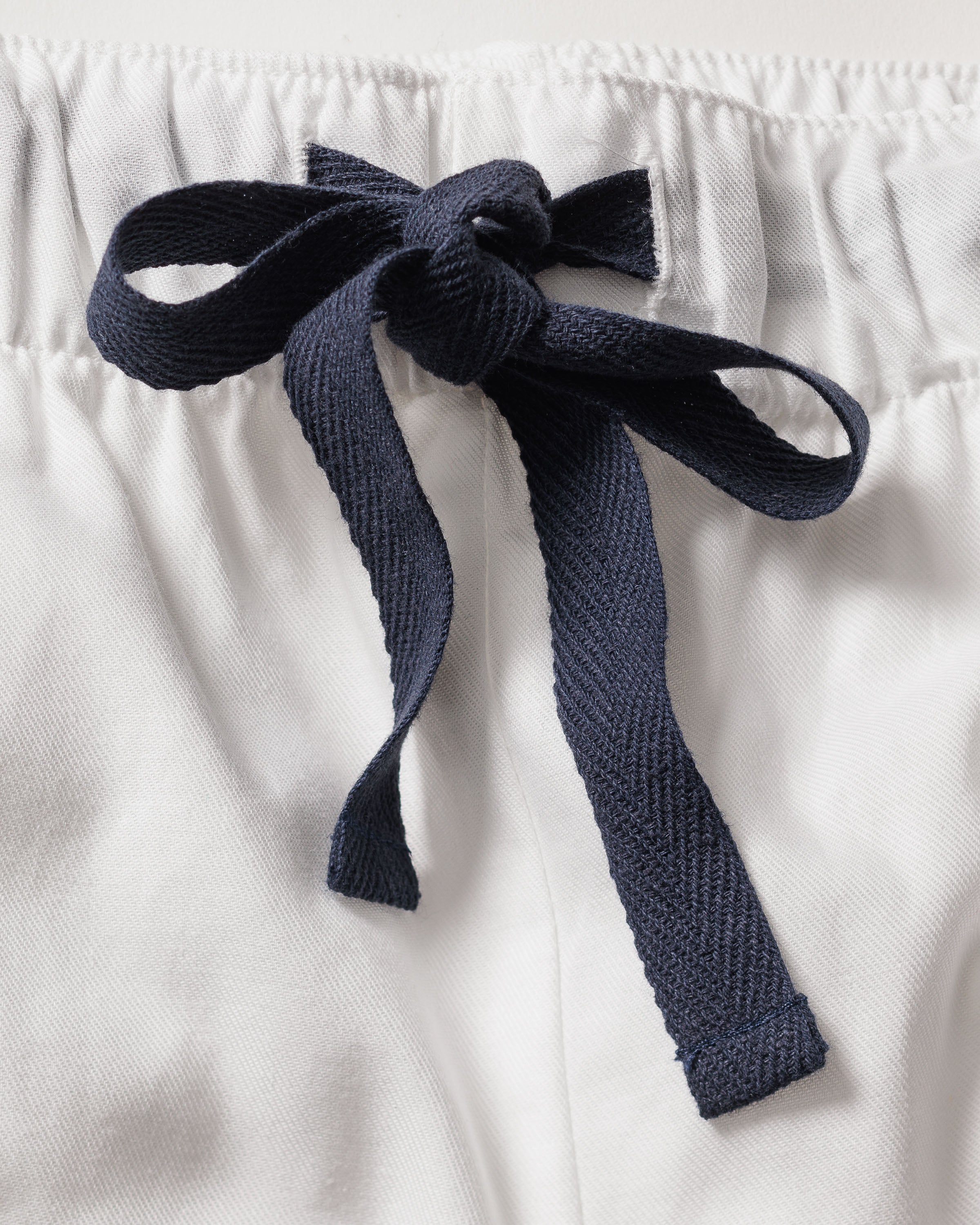 Close-up of Petite Plumes Womens Twill Pajama Set in White with Navy Piping, showcasing a cotton twill design with an elastic waistband and a black fabric bow at the center. The texture highlights its soft, moisture-wicking material.