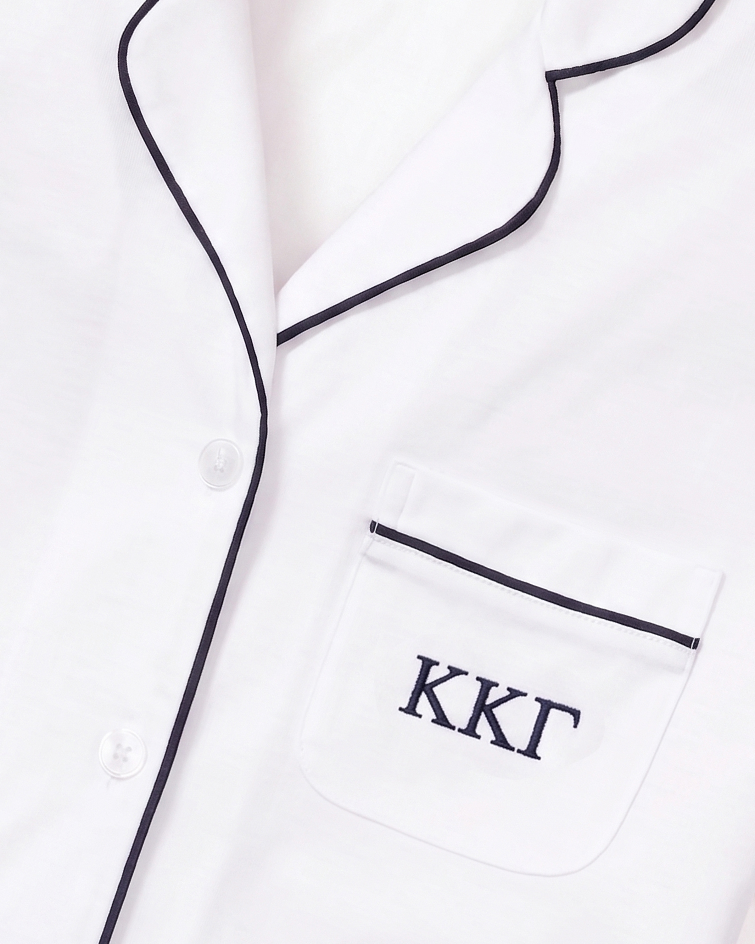 White Pima pajama top with navy piping and a pocket embroidered with the Greek letters ΚΚΓ. Two white buttons are visible along the front.