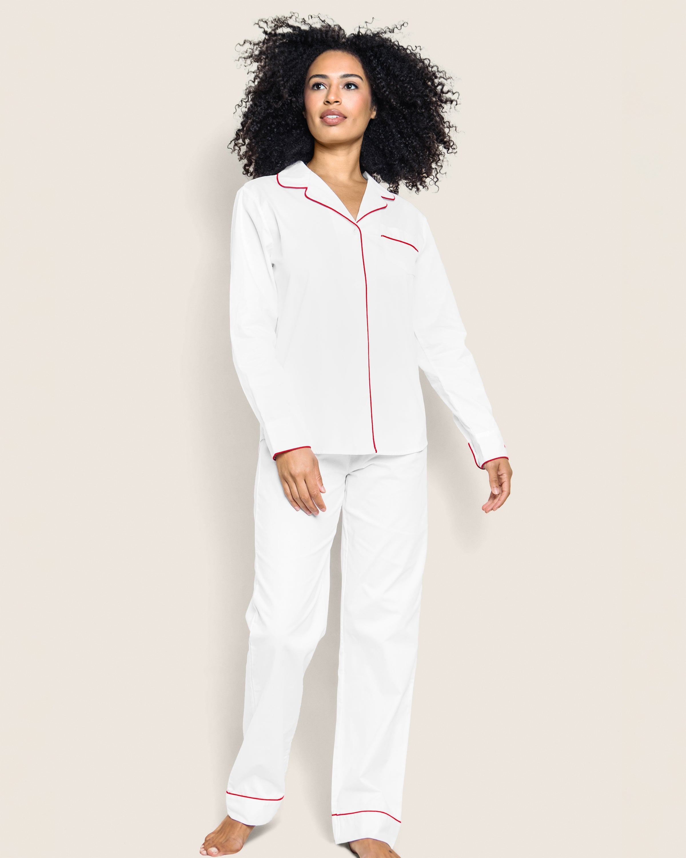 A person with curly hair is wearing Petite Plumes Womens Twill Pajama Set in White with Red Piping, featuring a collared long-sleeve shirt and long pants for style and moisture-wicking comfort. The background is plain light beige.