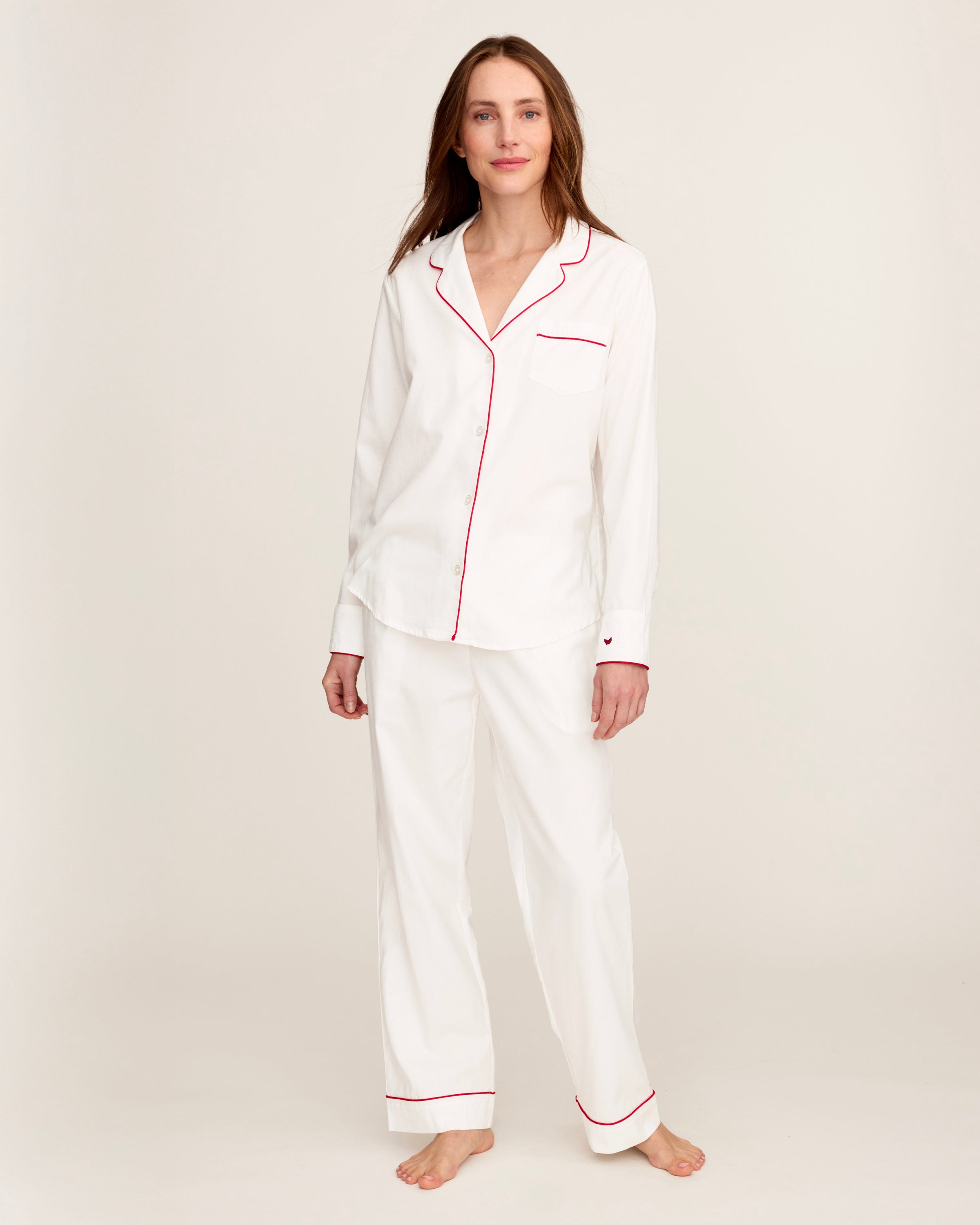 A woman stands barefoot in the Women's Twill Pajama Set in White with Red Piping against a plain, light background.