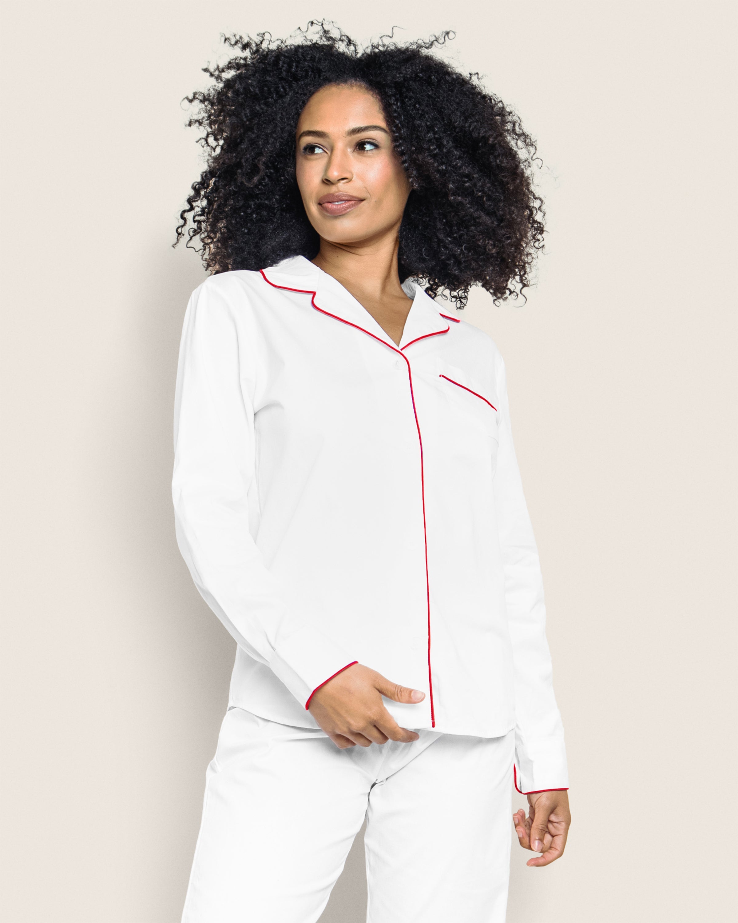 Someone with curly hair stands smiling slightly in a Womens Twill Pajama Set in White with Red Piping by Petite Plume, featuring red accents on the collar, pocket, and cuffs against a plain, light backdrop.