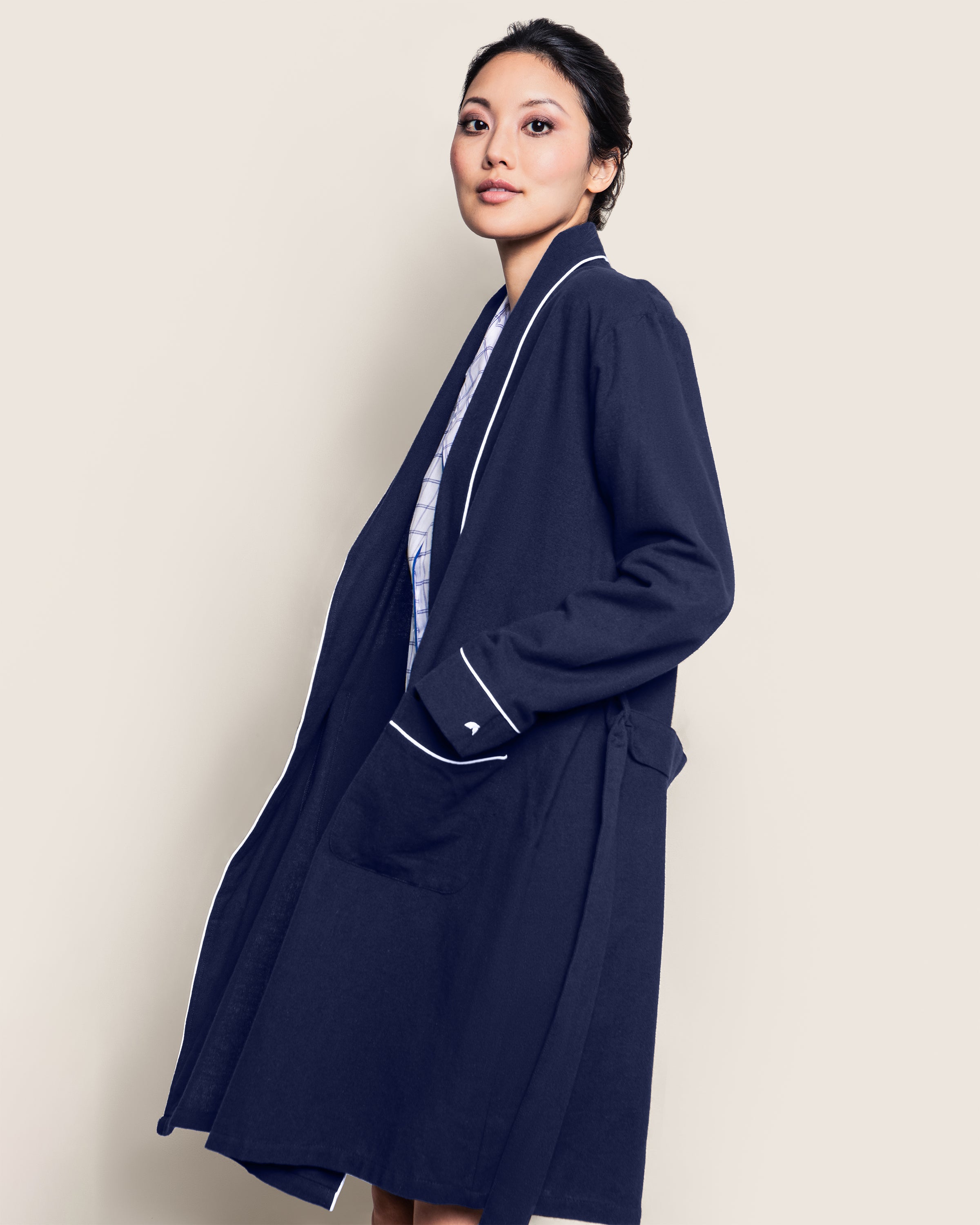 A woman in a relaxed pose glances over her shoulder, wearing a Petite Plume Womens Flannel Robe in Navy with white piping. Her hair is tied back as the 100% cotton twill fabric drapes effortlessly against a neutral background.