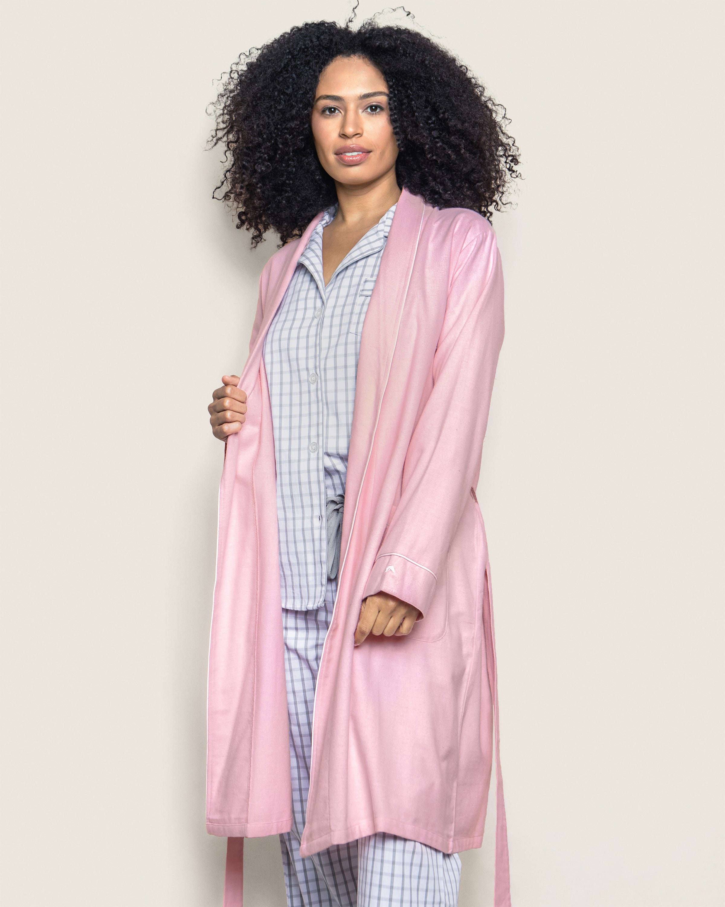 A person with curly hair confidently gazes at the camera wearing a Womens Flannel Robe in Pink by Petite Plume, open over 100% cotton checkered pajamas against a plain background.