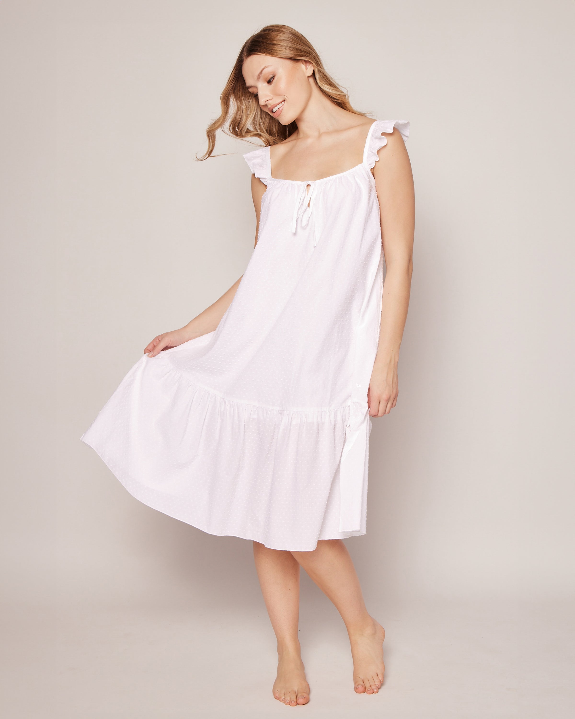 Barefoot, a person poses in Petite Plumes Womens Swiss Dots Celeste Nightgown in White. The sleeveless dress features ruffled straps and delicate cotton Swiss dots. Its flowing hem evokes the elegance of luxury sleepwear as they smile gently, looking downwards and holding the dresss side.