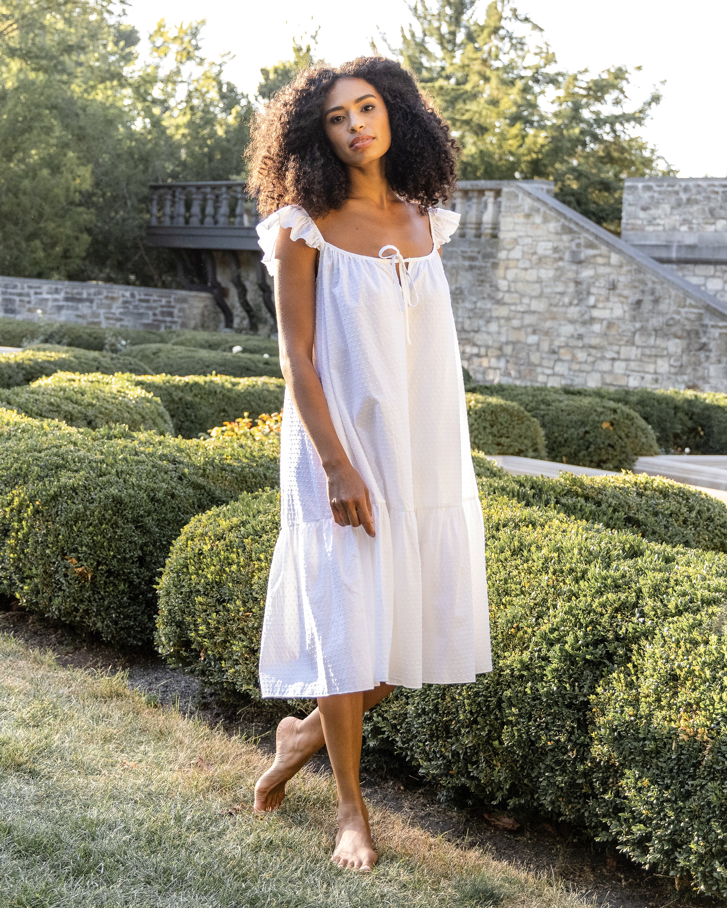 A barefoot woman with curly hair stands on grass in a garden wearing Petite Plumes Womens Swiss Dots Celeste Nightgown in white, featuring ruffled straps, amid manicured hedges and a stone wall, all gently illuminated by soft sunlight.