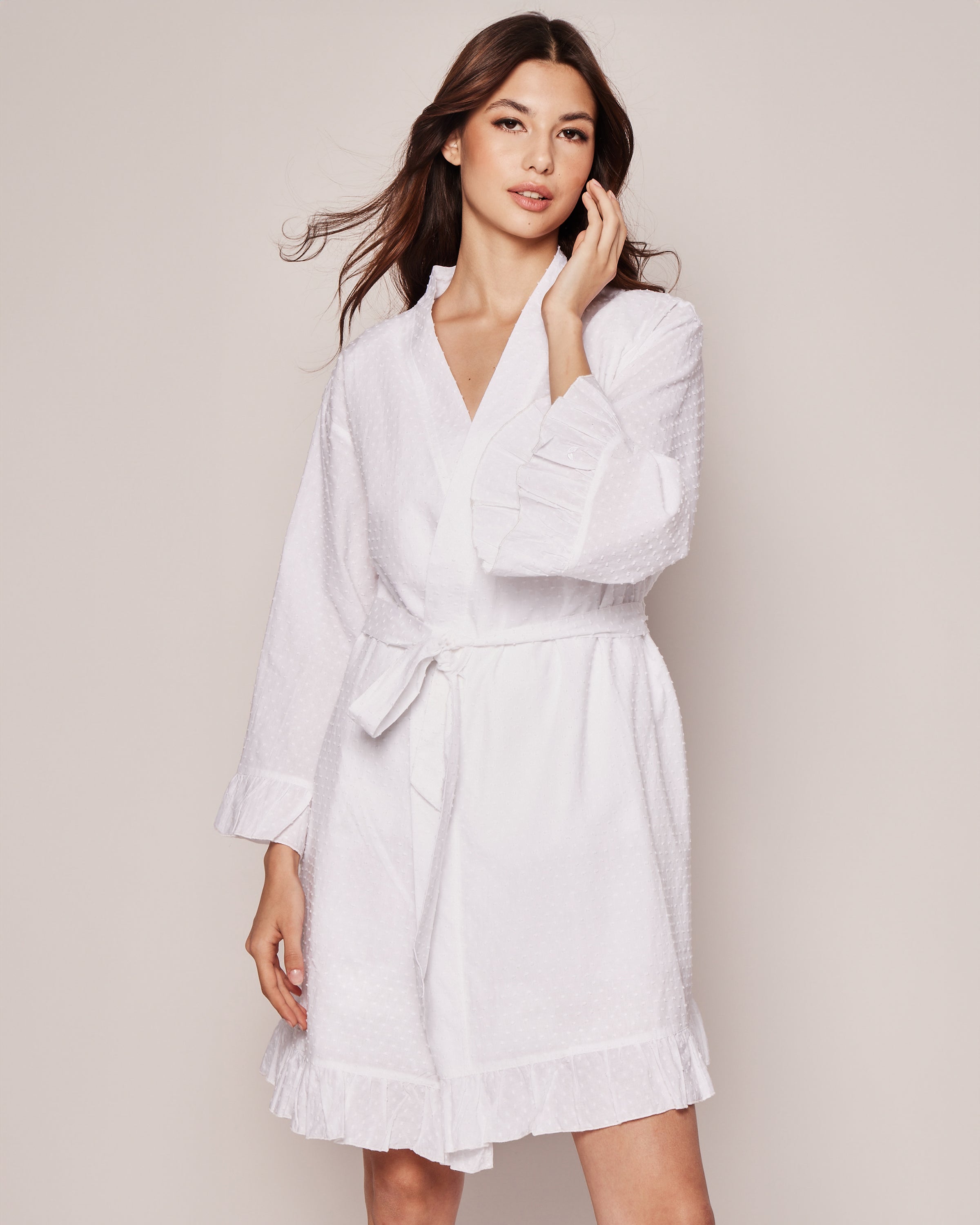 A woman models the Petite Plume Womens Swiss Dots Robe in white cotton, showcasing its ruffled cuffs and hem. She stands against a light background, touching her hair.