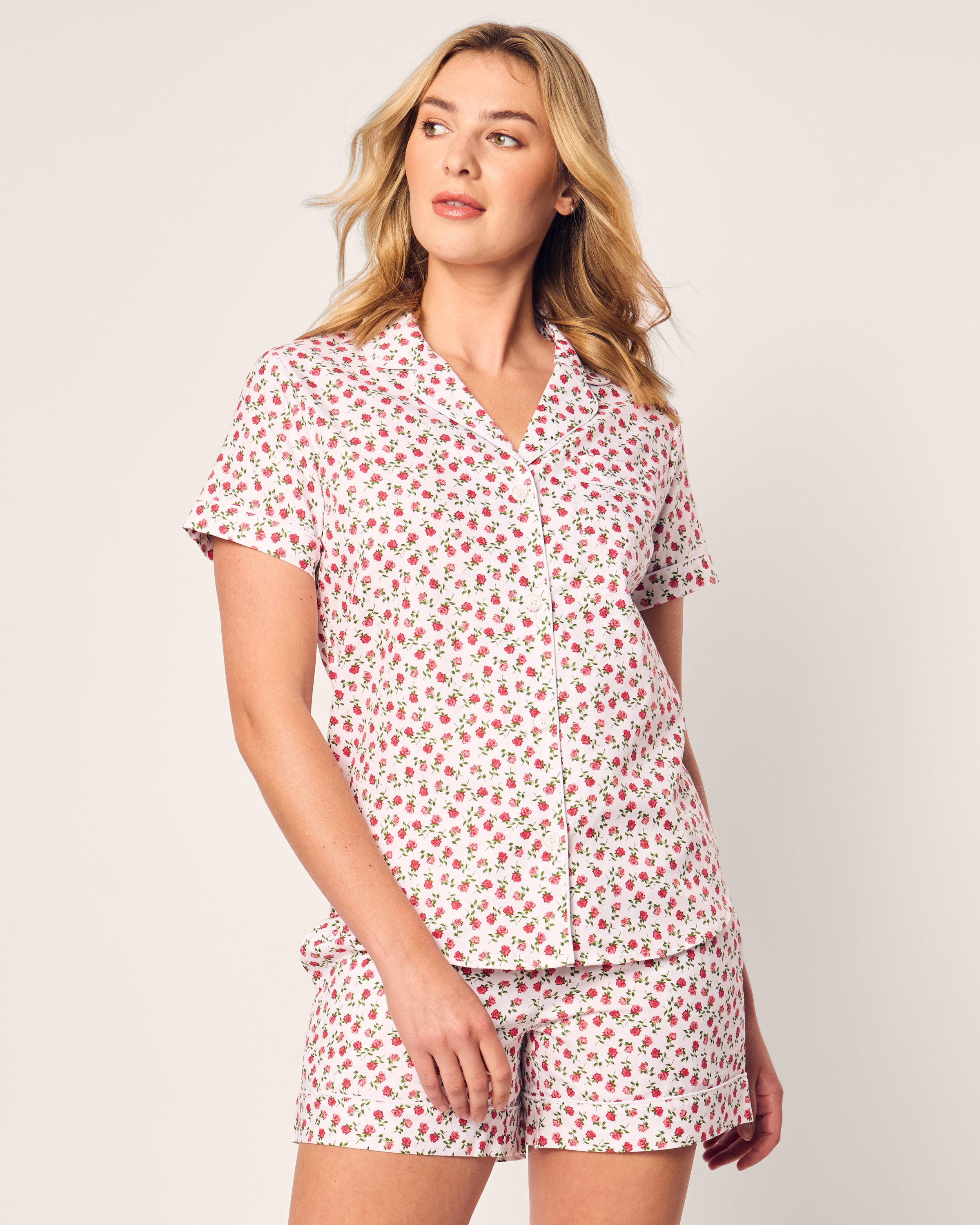 A person in Petite Plumes Womens Twill Short Sleeve Short Set in Petite Petals, featuring a white base with pink florals, stands against a plain background, gazing to the side.