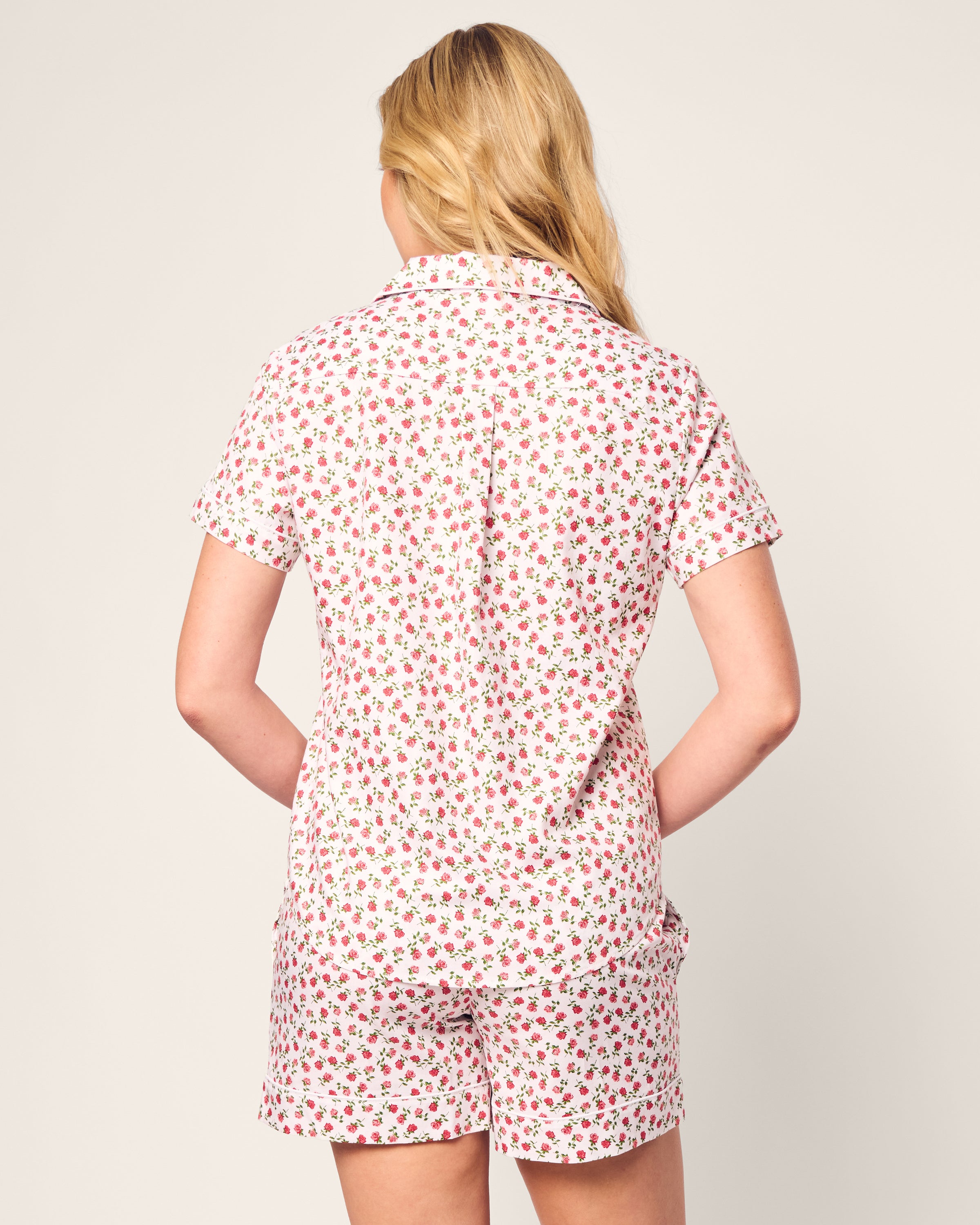 A person with long blonde hair is seen from the back, wearing Petite Plumes Womens Twill Short Sleeve Short Set in Petite Petals. The luxurious outfit is white with a small red floral pattern, standing out elegantly against the plain off-white background.