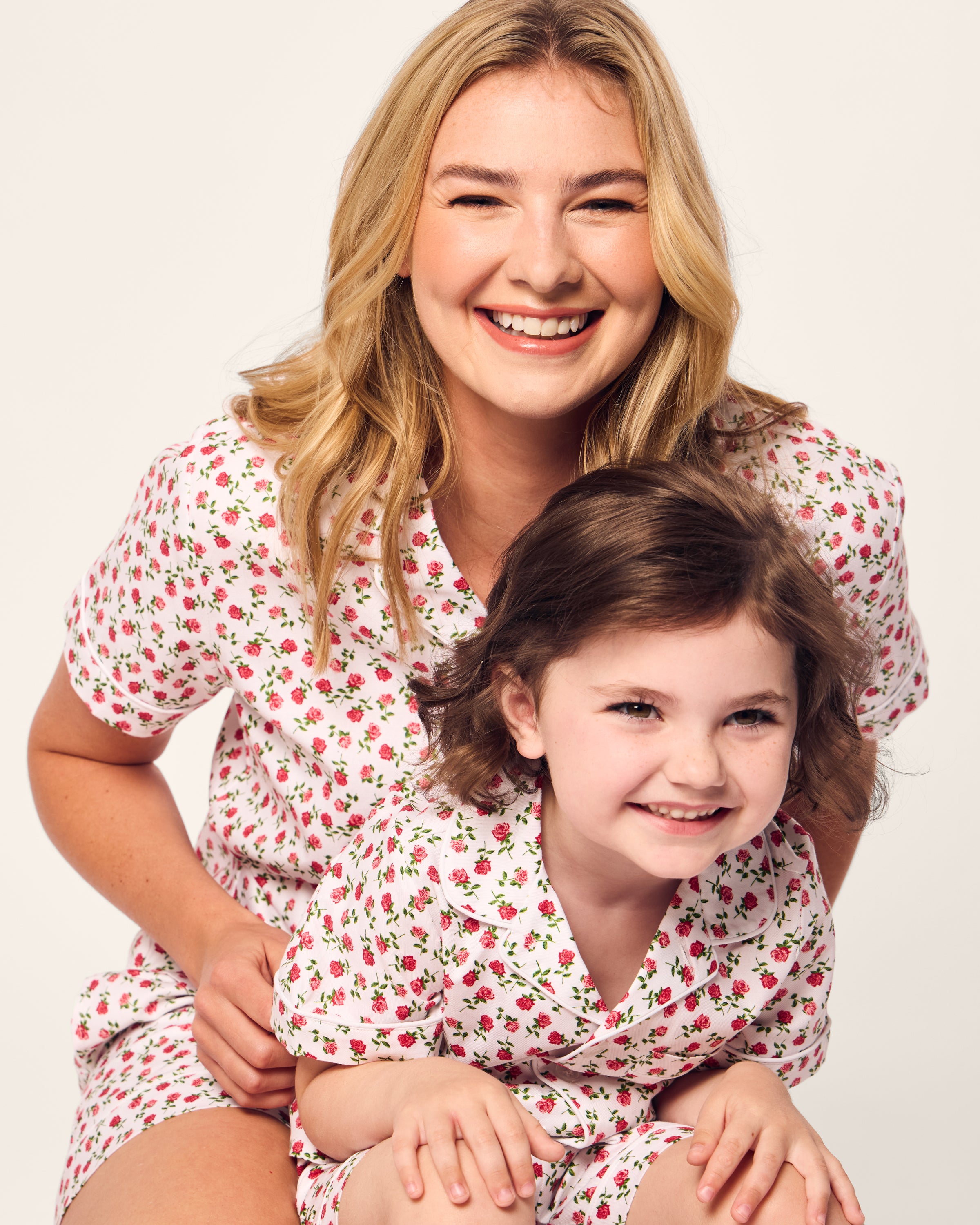 A smiling woman and a young girl in matching Petite Plume Womens Twill Short Sleeve Short Sets in Petite Petals pose together against a light background. The woman sits slightly behind the girl, embracing her around the shoulders, and both are looking towards the camera.