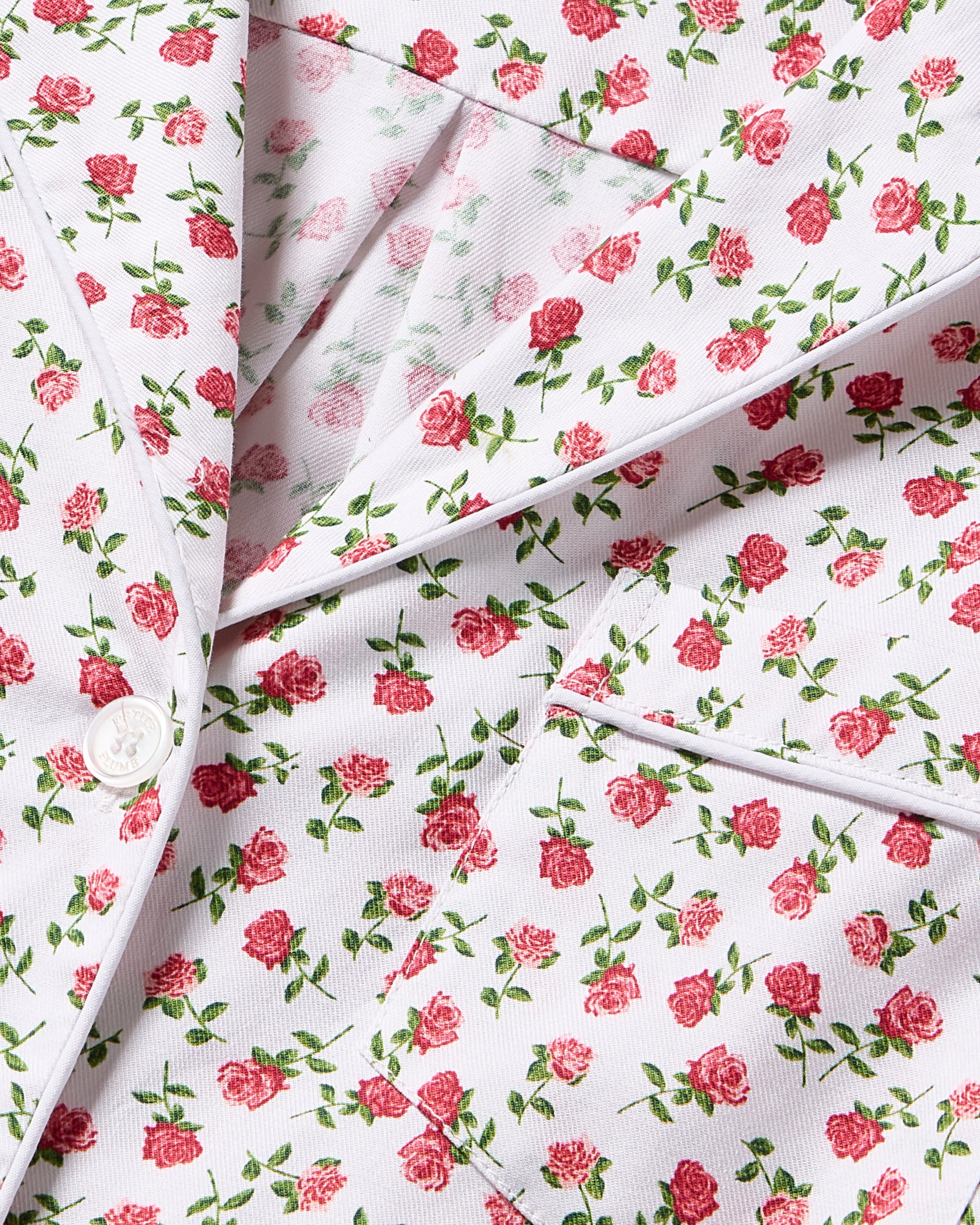 A close-up of Womens Twill Short Sleeve Short Set in Petite Petals by Petite Plume shows its elegant floral design with pink roses on a white background, featuring visible lapels, a button, and a pocket.