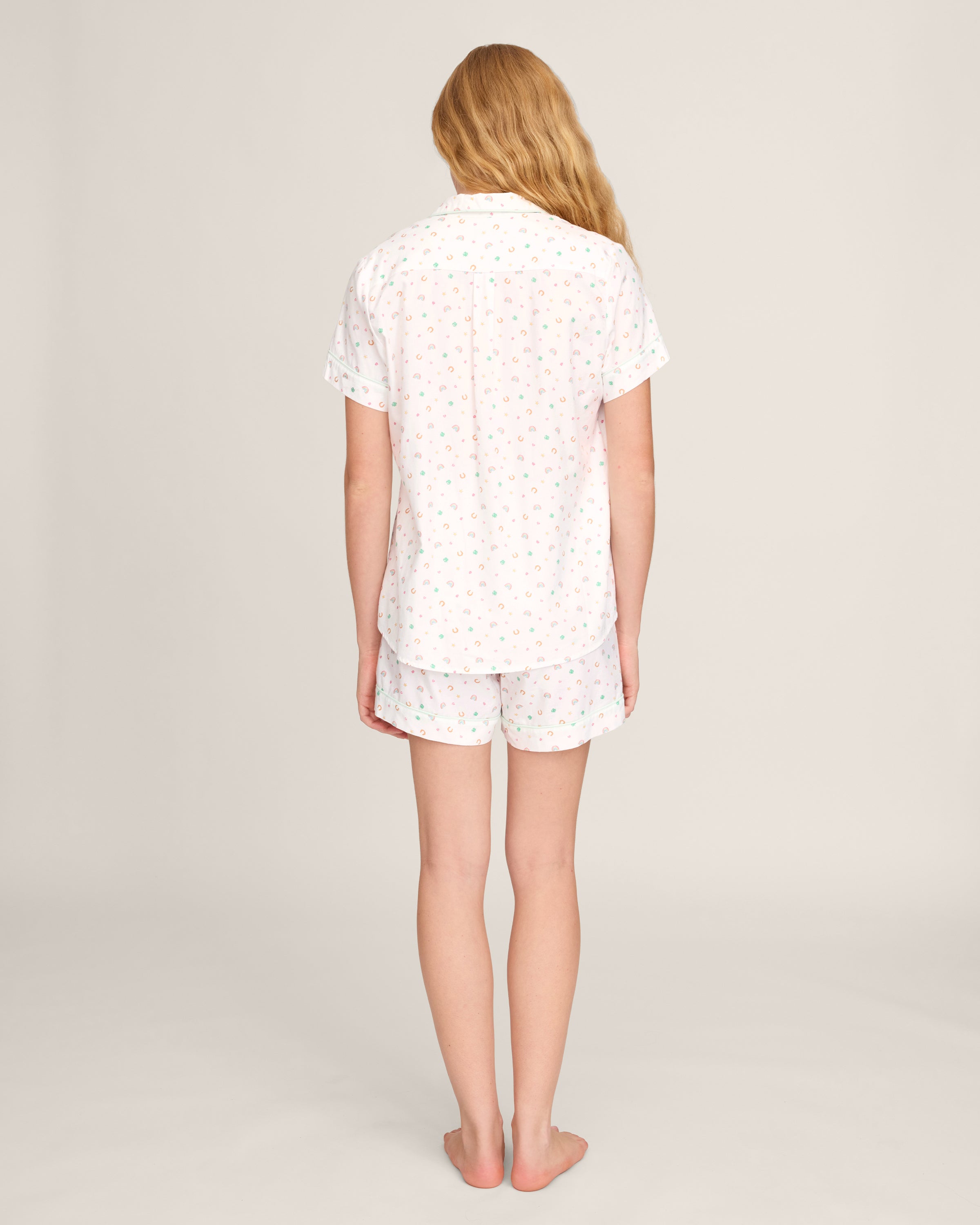 A woman with long, wavy blonde hair wears the Women's Twill Short Sleeve Short Set in Good Luck Charms, featuring a multicolored pattern, standing against a plain light background.