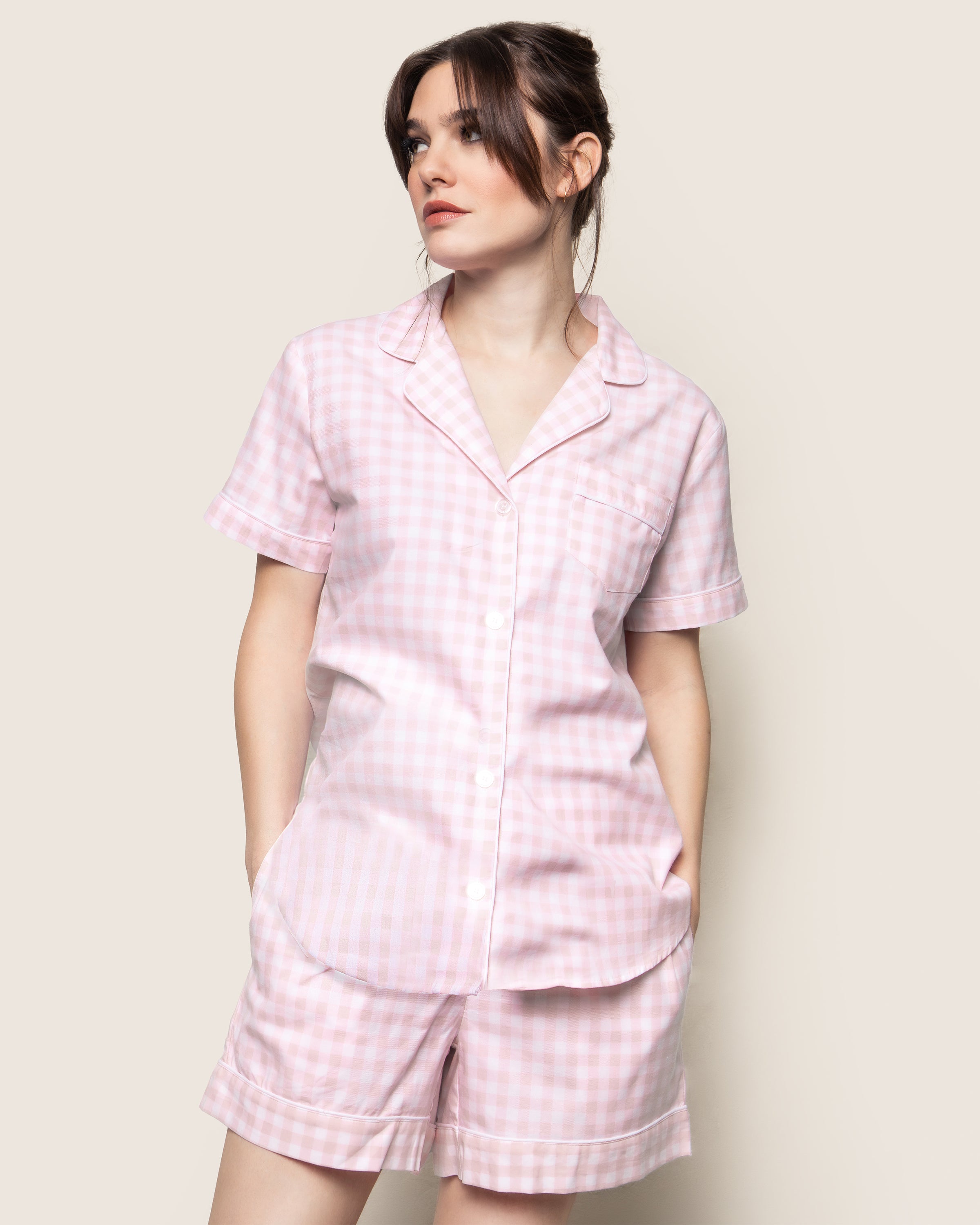 A person models Petite Plumes Womens Twill Pajama Short Set in Pink Gingham. The soft cotton set features a short-sleeved button-up shirt and matching shorts, blending casual style with cozy comfort against a plain background.