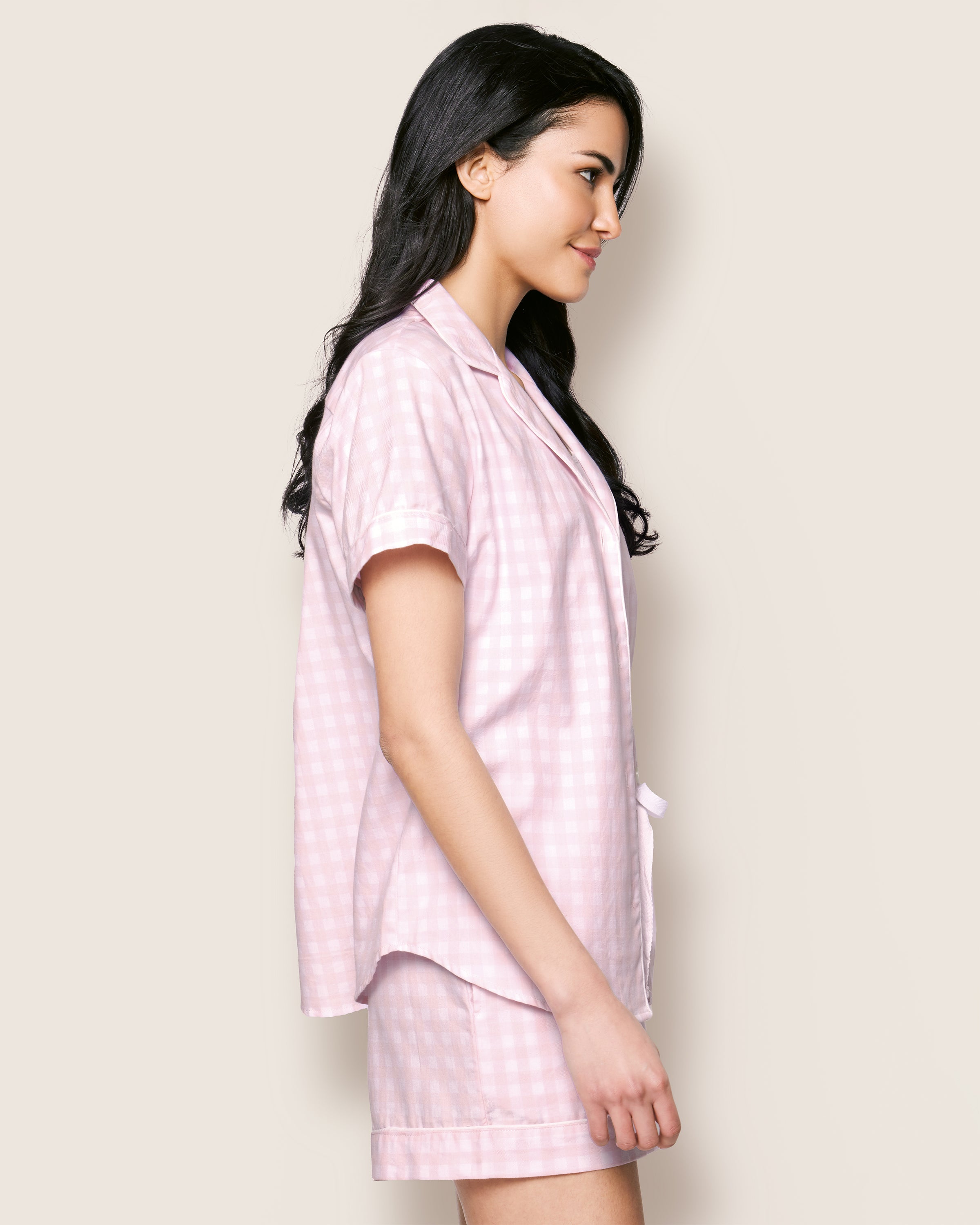 A person with long dark hair stands in profile, wearing Petite Plumes Womens Twill Pajama Short Set in Pink Gingham, featuring a cotton short-sleeve button-up shirt and shorts, set against a plain light background.