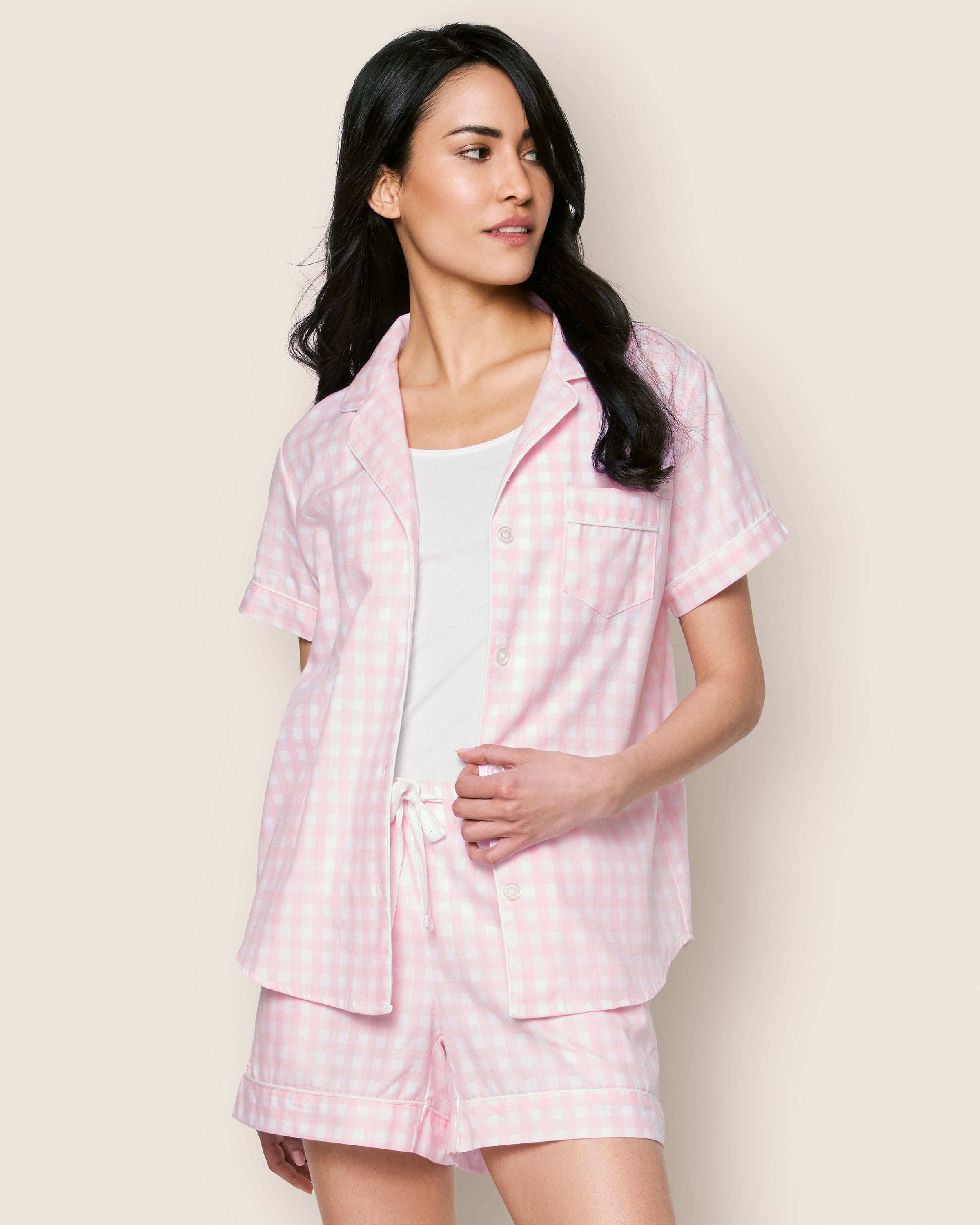 A woman with long dark hair poses against a plain background, elegantly dressed in Petite Plumes Womens Twill Pajama Short Set in Pink Gingham. The short sleeves, shorts, and open collar create an effortlessly chic look as she gazes thoughtfully to the side.