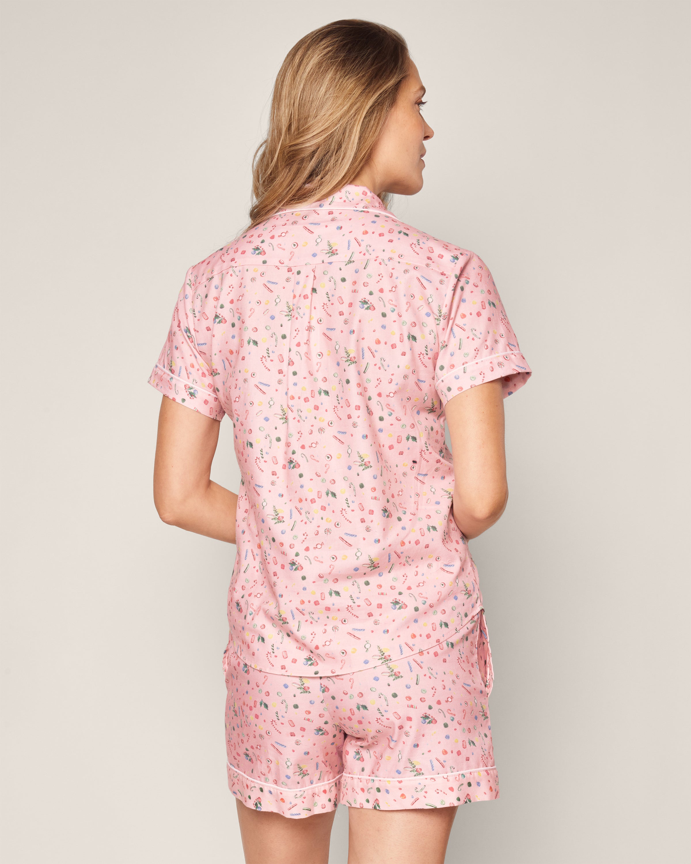 A person with long hair wears Petite Plumes Womens Twill Short Sleeve Short Set in Vintage Sweets, a light pink 100% cotton pajama set with a colorful pattern. The short-sleeved button-up top and shorts are modeled from the back, highlighting the charming design.