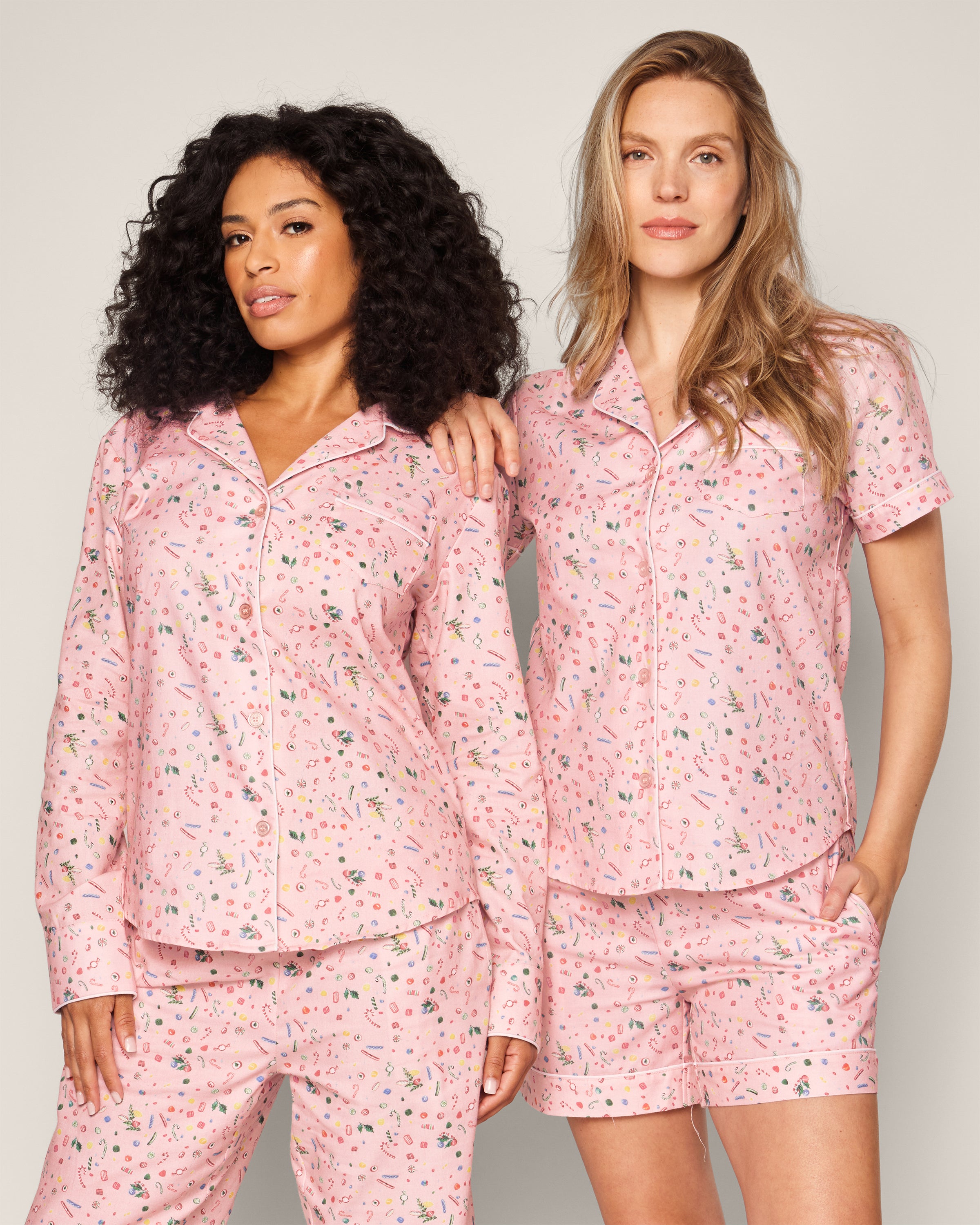 Two people wear matching pink pajama sets with small patterns. The left person has long curly hair in a long-sleeve set, while the right one with straight hair wears the Petite Plume Womens Twill Short Sleeve Short Set in Vintage Sweets. Both are crafted from 100% cotton.
