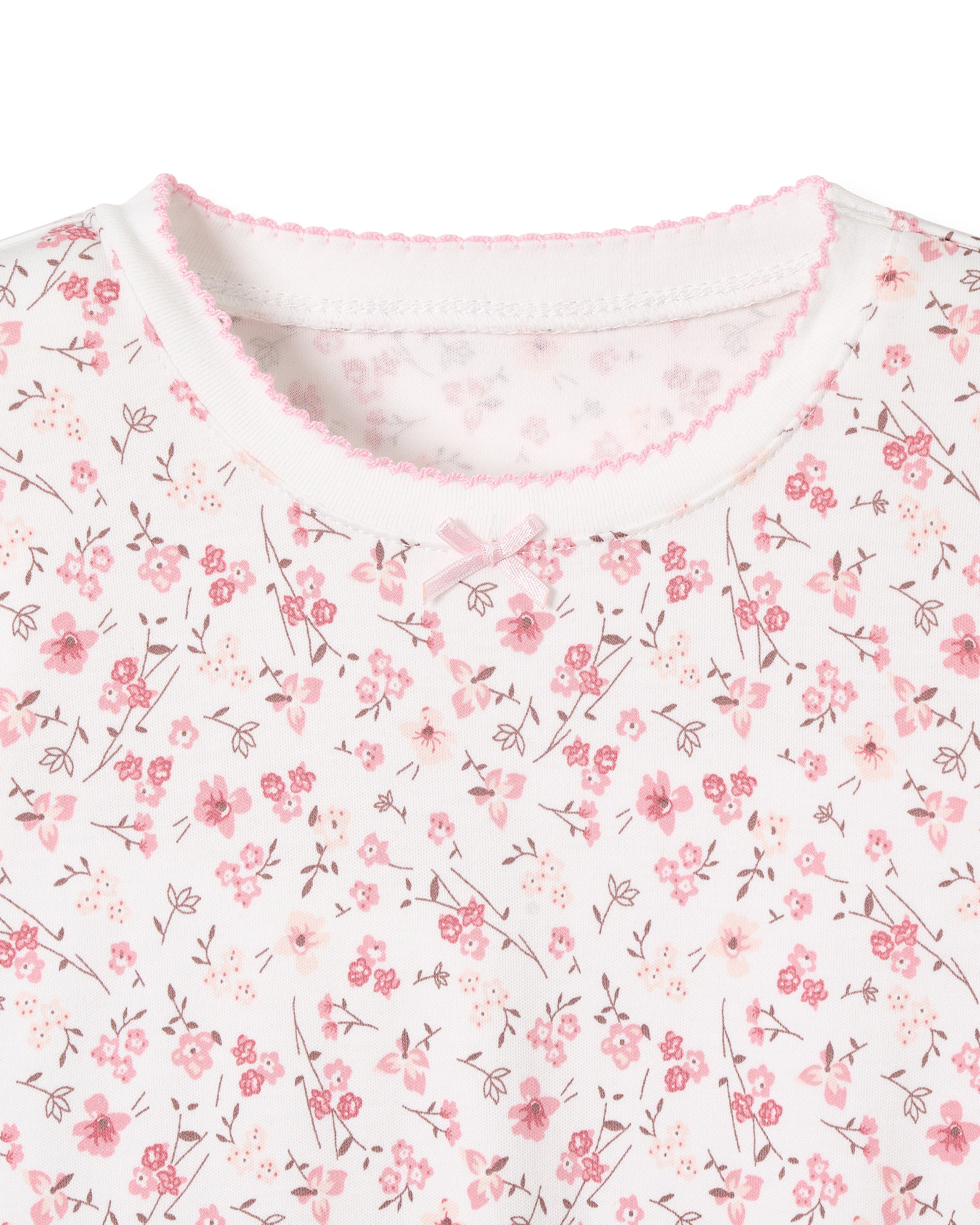 Close-up of Kids Pima Snug Fit Pajama Short Set in Dorset Floral by Petite Plume: white fabric decorated with pink flowers and branches, featuring a round neckline, pink zigzag stitching, and a tiny bow at the center.
