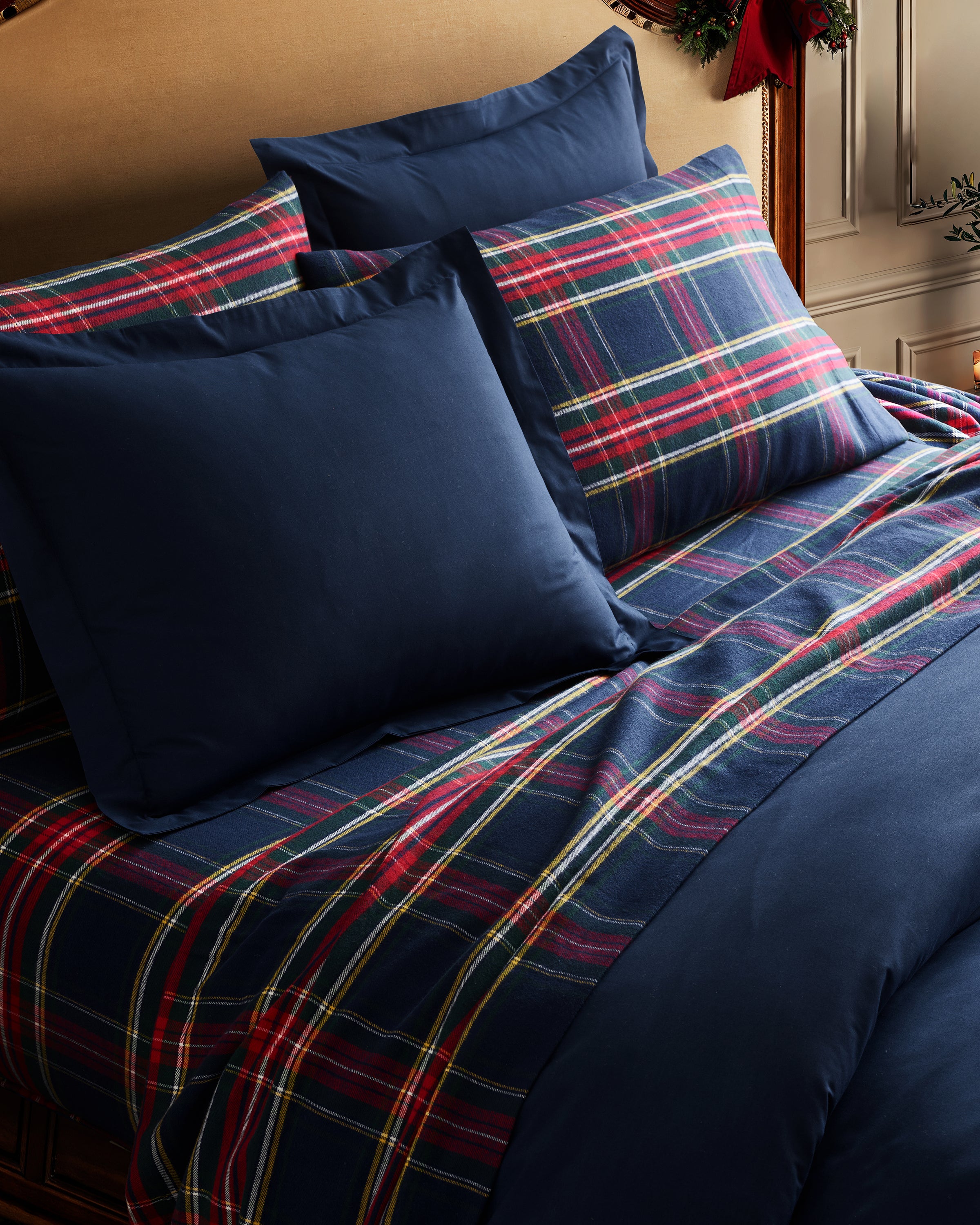 A neatly made bed showcases Petite Plumes Flannel Sheets in Windsor Tartan, featuring navy blue and red plaid cotton brushed flannel bedding with matching pillows. Set against a wooden headboard, the scene hints at a cozy, classic bedroom aesthetic.
