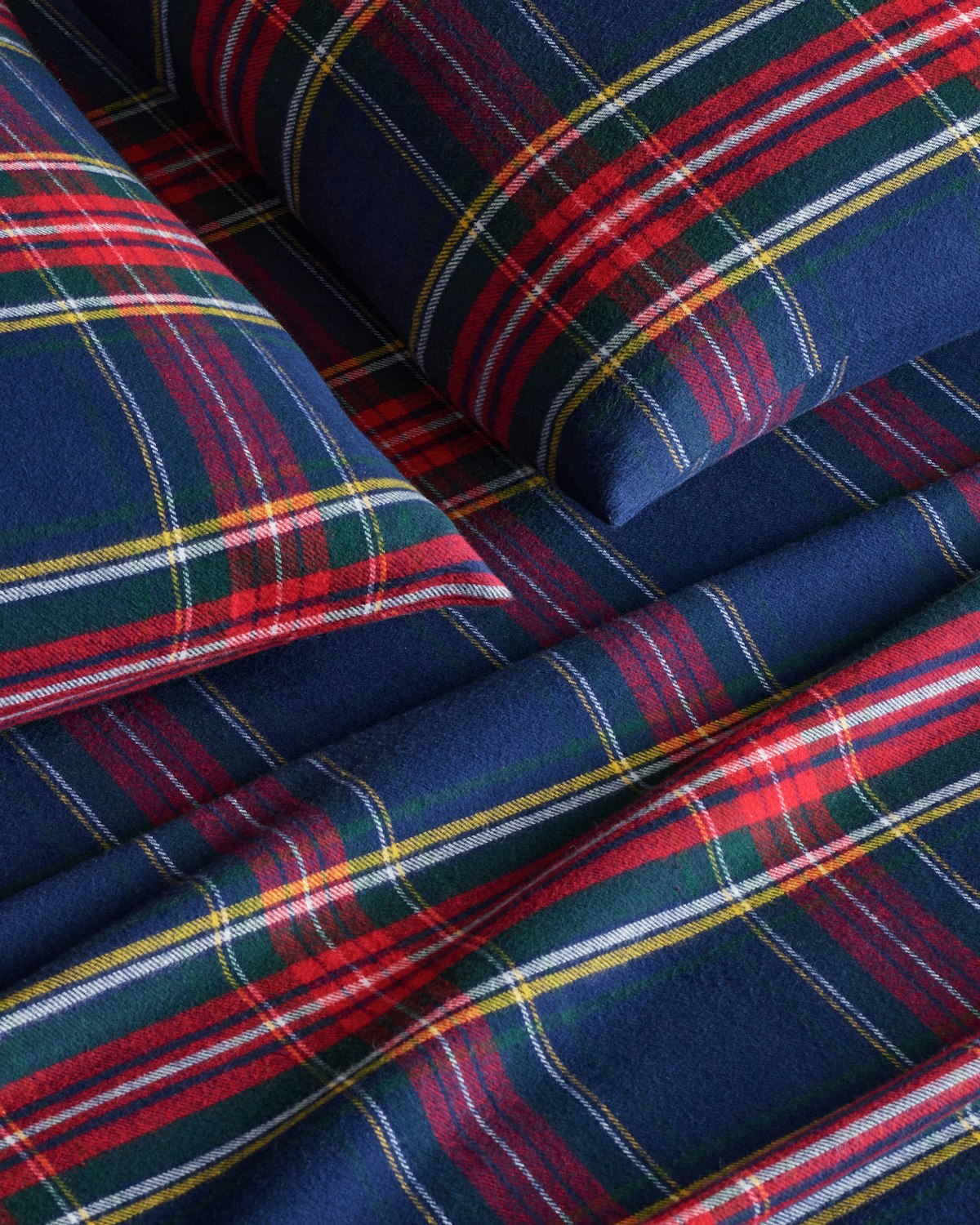 Close-up of a bed with Petite Plumes Flannel Sheets in Windsor Tartan, featuring cozy plaid cotton brushed flannel sheets and pillowcases in blue, red, green, and white stripes.