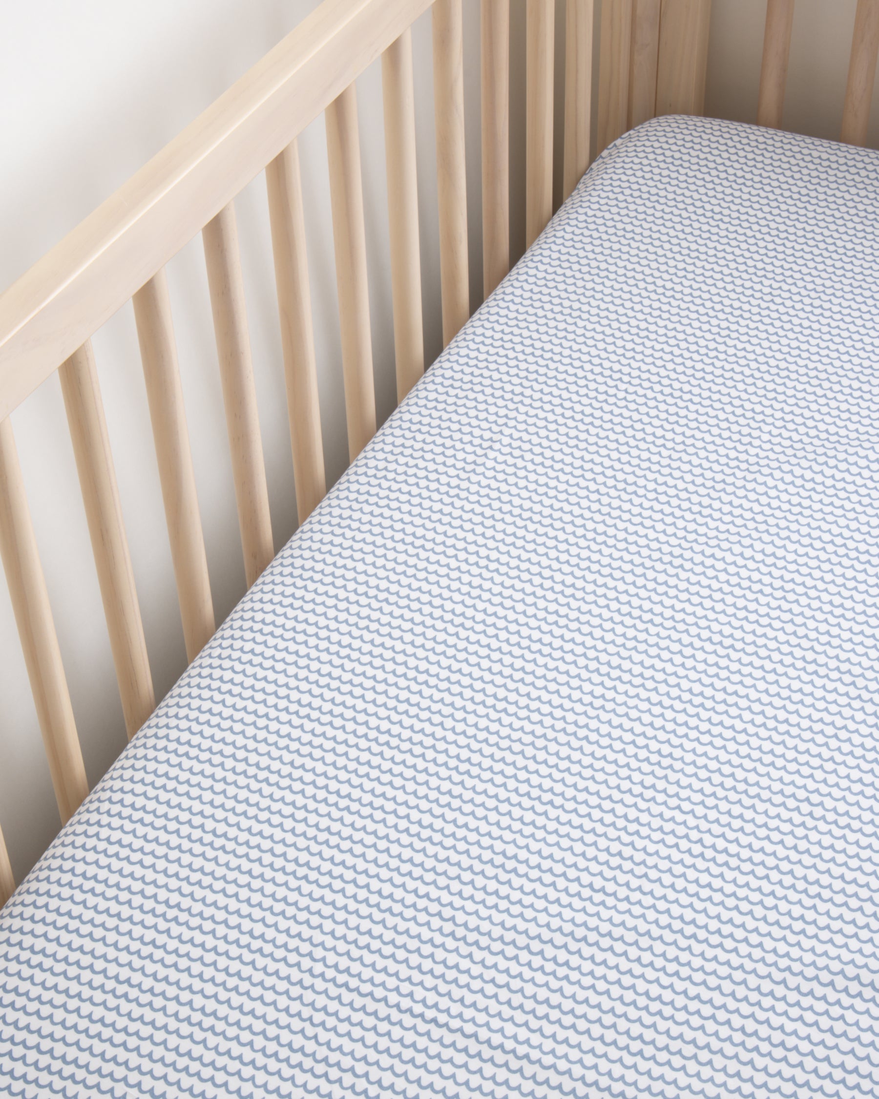 A wooden crib showcases an ultra-soft cotton percale mattress topped with Petite Plumes Luxe Premium La Mer Crib Sheet, a breathable white cover with a subtle blue scallop pattern, set against light wood slats and a white wall.
