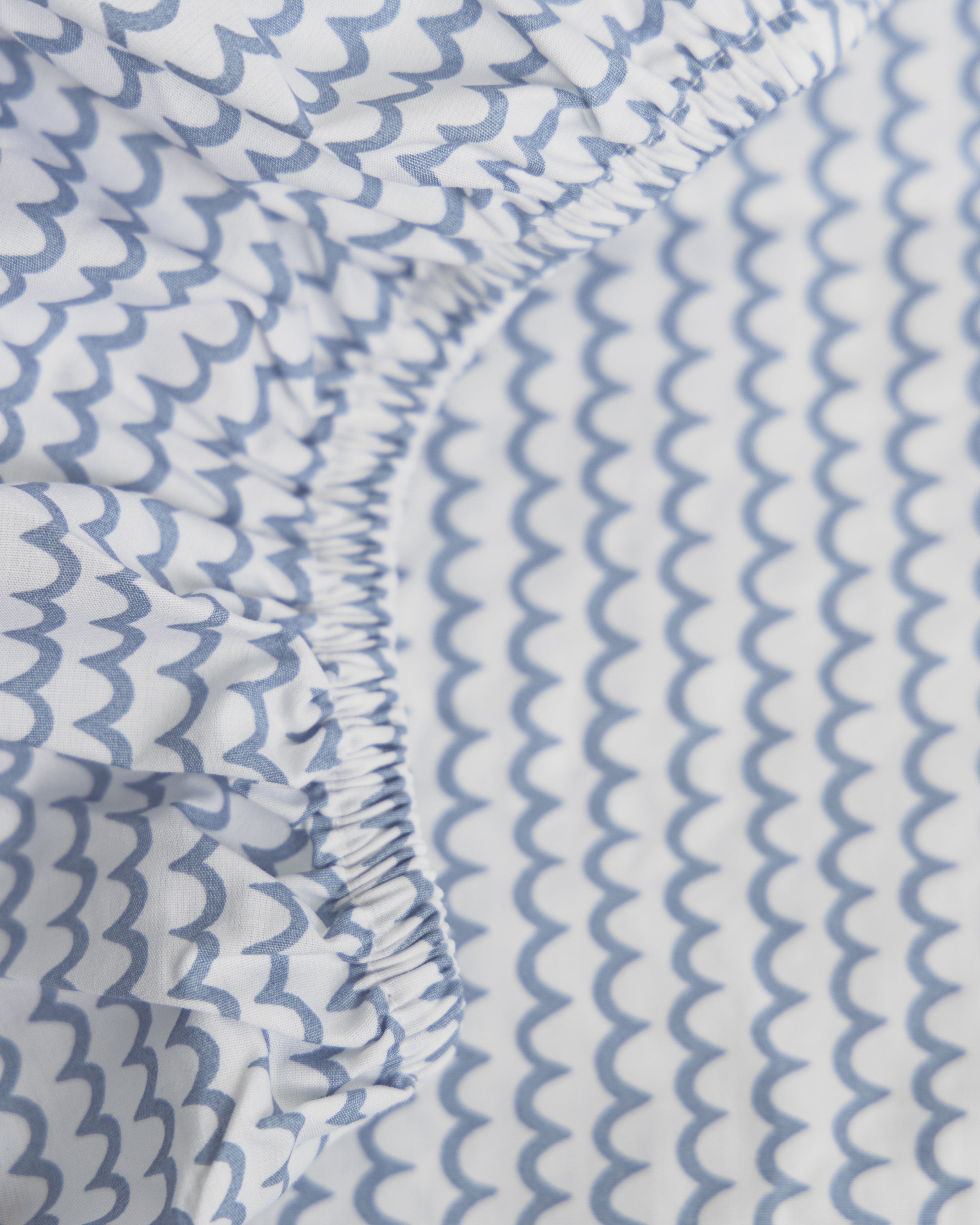 Close-up of Petite Plumes Luxe Premium La Mer Crib Sheet featuring a light blue scalloped pattern on white. The breathable, ultra-soft cotton percale fabric is gathered with elastic for a ruched effect, ideal for coziness.