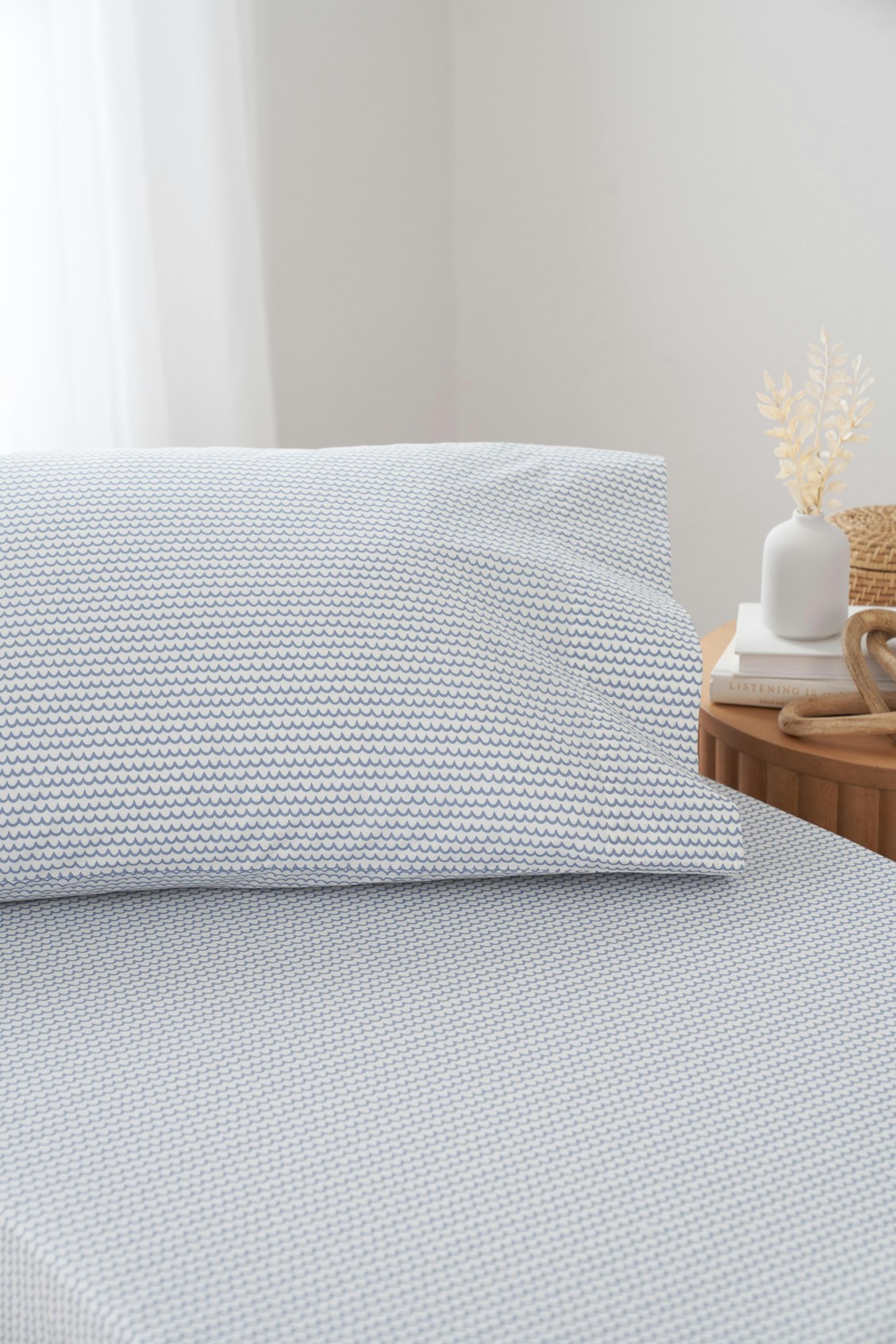 A neatly made bed showcases Luxe Premium Cotton Bed Sheets in Timeless Toile by Petite Plume, paired with pillowcases. A bedside table holds a white vase with dried plants, stacked books, and a woven basket. Soft natural light filters through a sheer curtain.