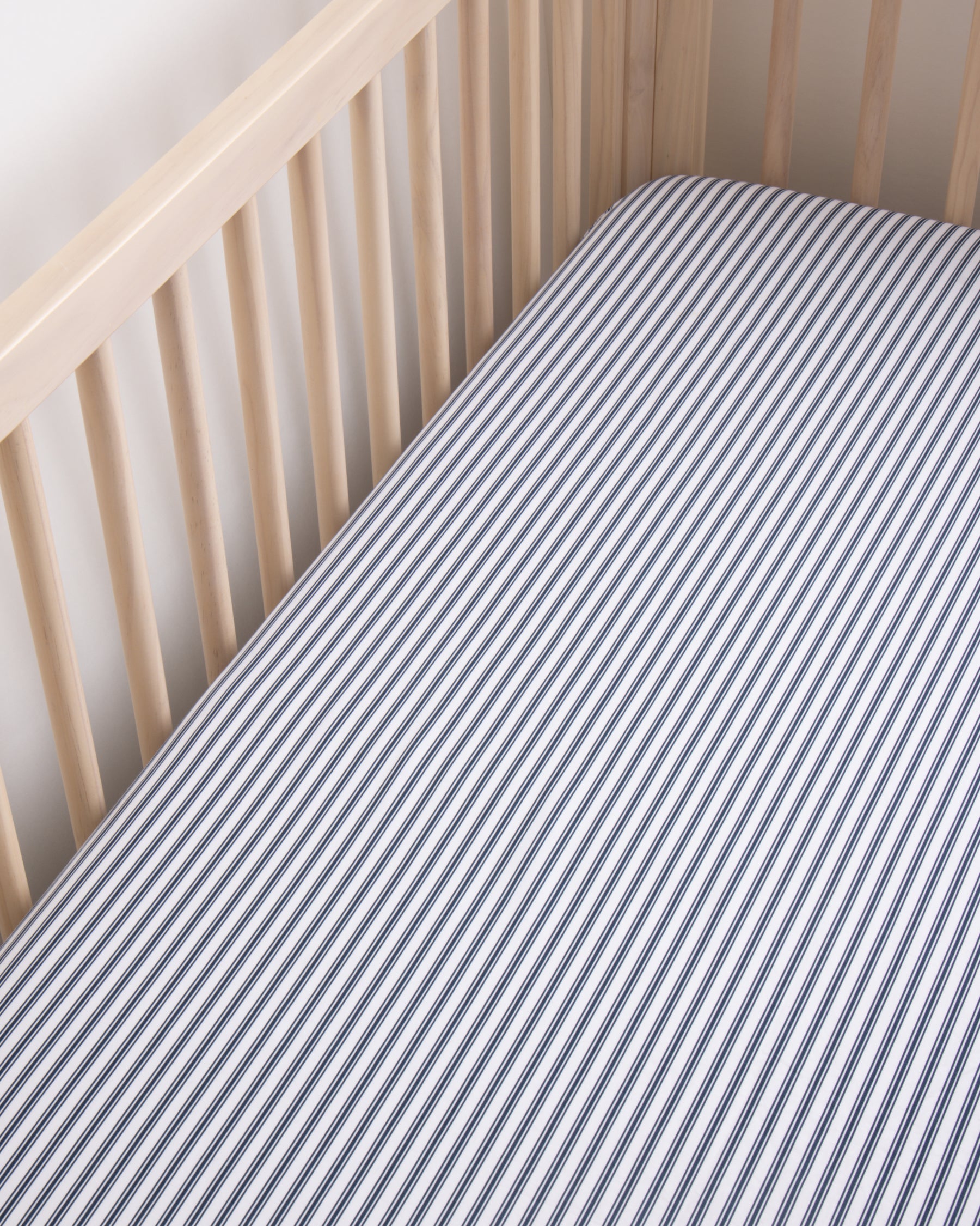 A light wood crib fitted with Petite Plumes Luxe Premium Cotton Crib Sheet in Navy French Ticking, featuring breathable, ultra-soft cotton percale in blue and white stripes. The design showcases a simple and clean nursery bedding look from above.