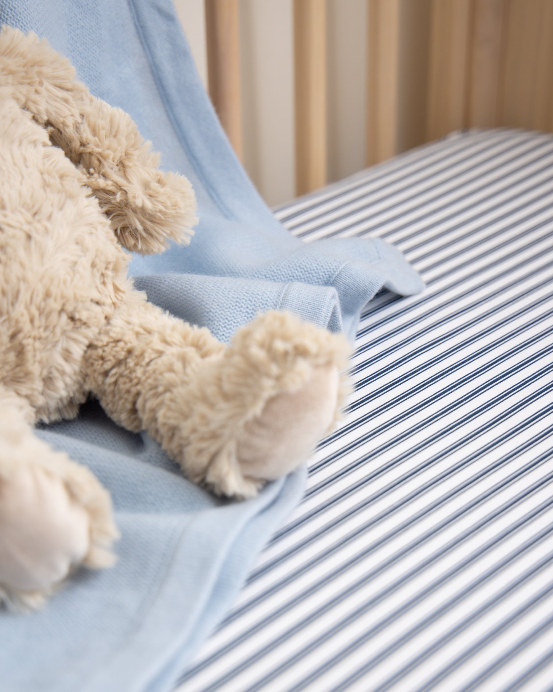 A close-up of a teddy bear on the Luxe Premium Cotton Crib Sheet in Navy French Ticking by Petite Plume is shown. A soft, light blue blanket drapes over the wooden railing, enhancing the cozy nursery setting with its ultra-soft texture.