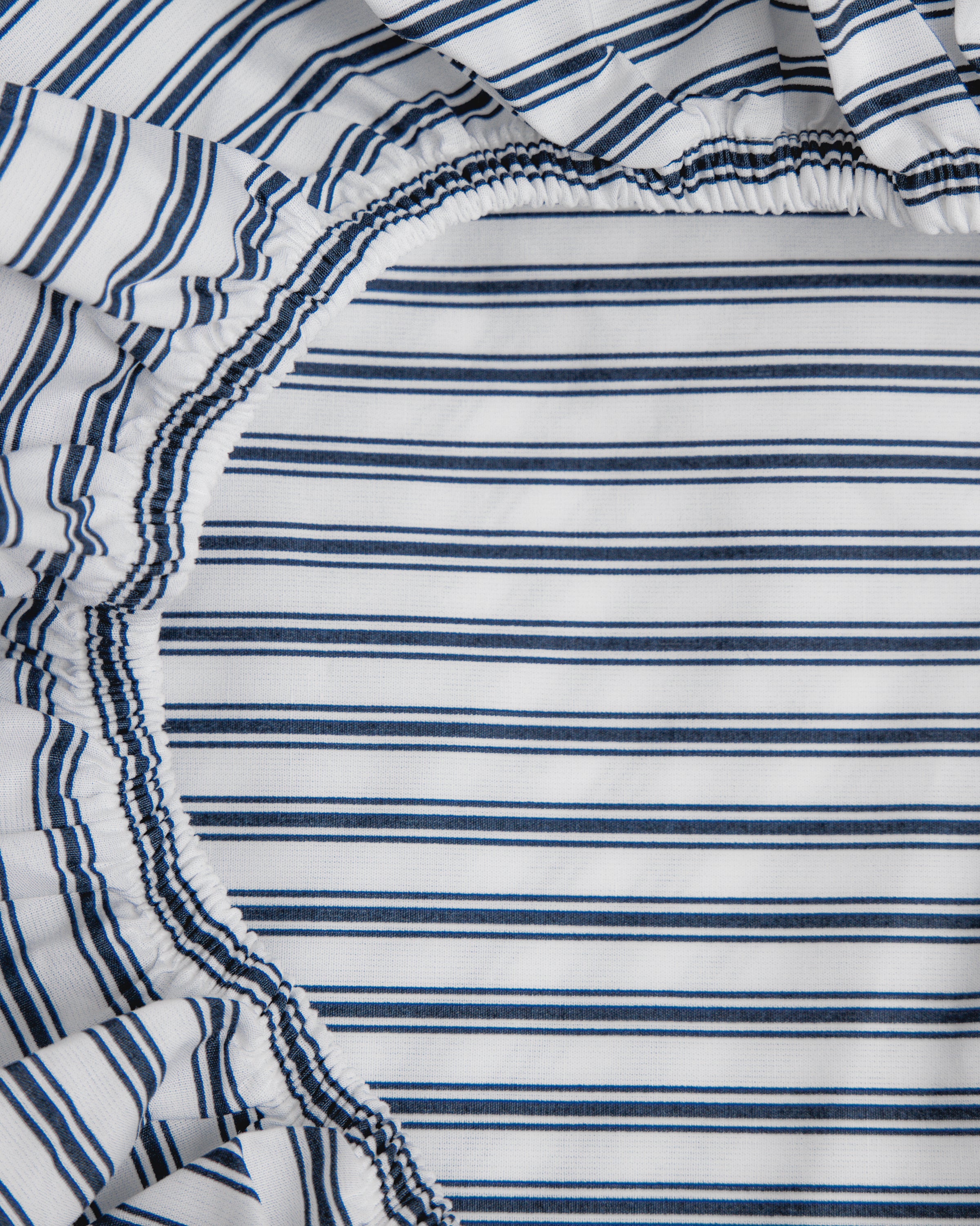 A close-up of Petite Plumes Luxe Premium Cotton Crib Sheet in Navy French Ticking shows breathable cotton percale with thin blue and white stripes, gathered with elastic into a circular shape at the corner.
