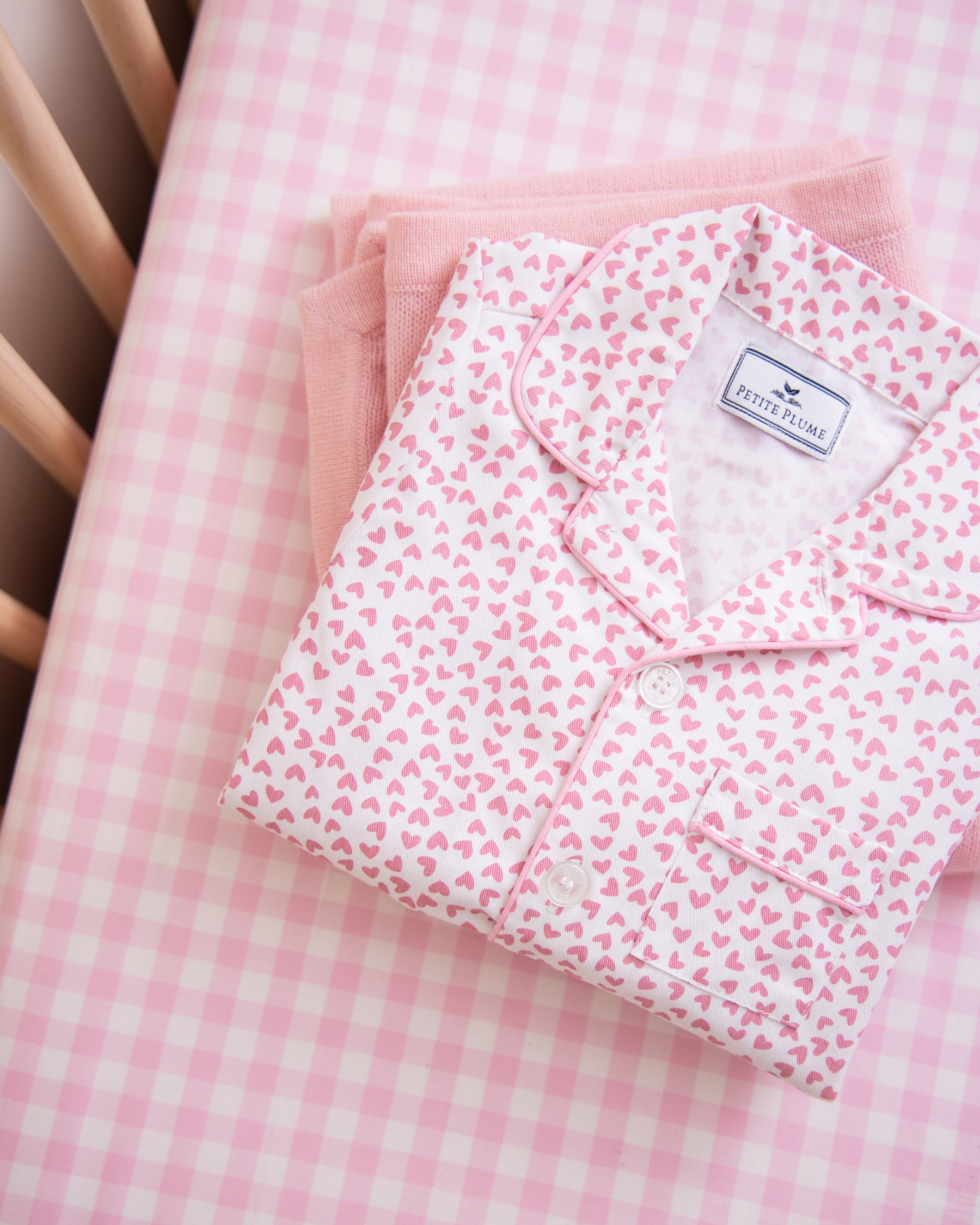 Folded pink pajamas with white hearts lie on a plush Luxe Premium Cotton Pink Gingham Crib Sheet by Petite Plume, along with a neatly folded pink towel. The beige crib railing peeks from the left side.