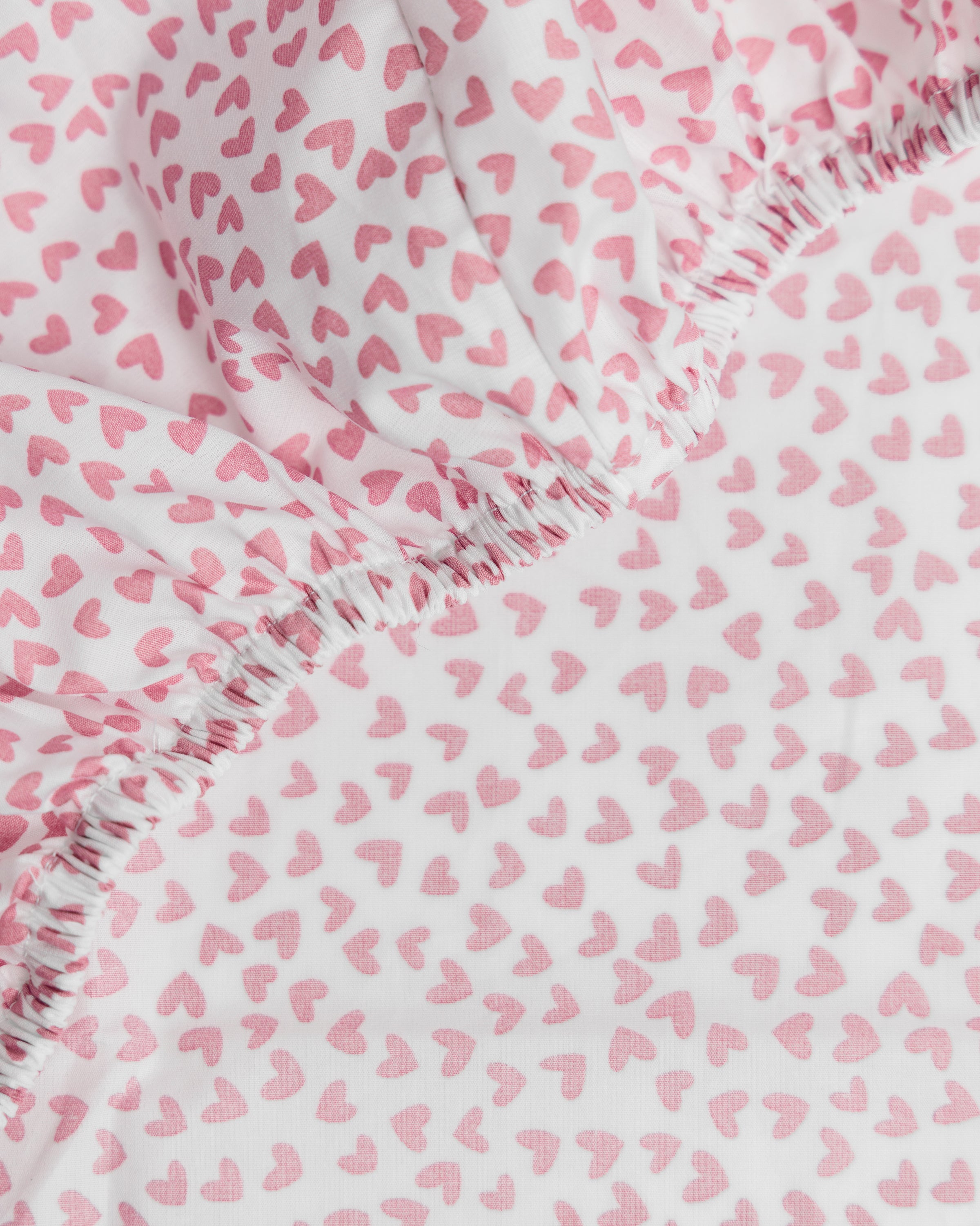 Close-up of the Luxe Premium Sweethearts Crib Sheet by Petite Plume, showcasing ultra-soft fabric with pink heart patterns on a white background. Made from breathable cotton percale with elastic edges, its playful design adds a cheerful touch to any nursery.