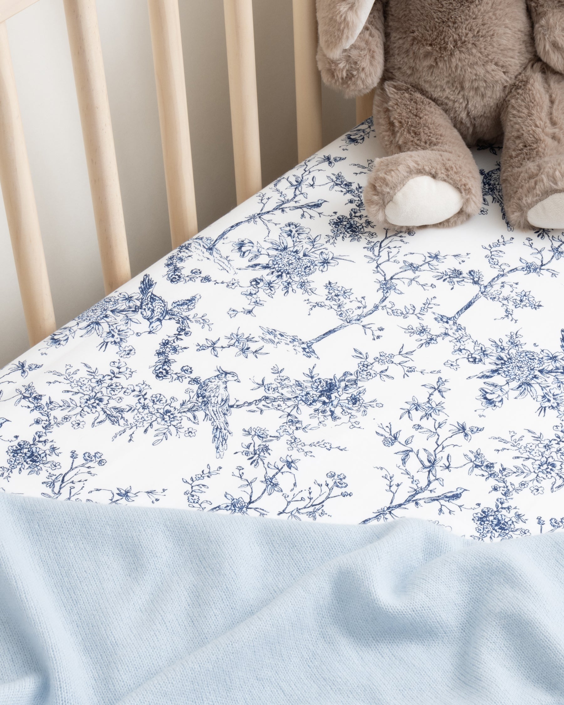 A crib with a Luxe Premium Cotton sheet by Petite Plume, showcasing a blue floral Timeless Toile pattern. A plush brown rabbit toy is seen in one corner with a light blue blanket partially showing at the bottom of the image.
