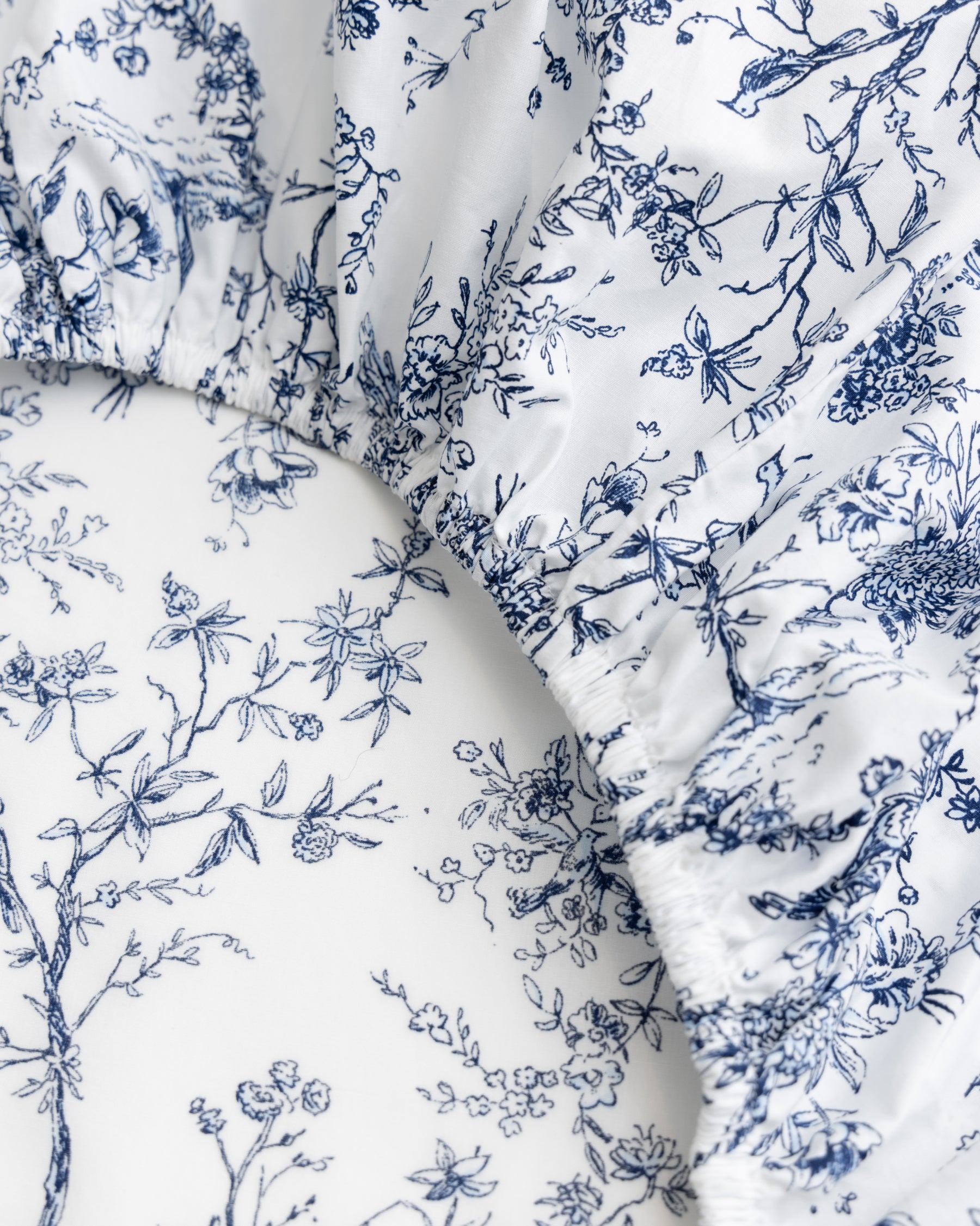 Close-up of Luxe Premium Cotton Crib Sheets in Timeless Toile by Petite Plume features ultra-soft fabric with intricate blue floral and vine patterns on white. Its gathered, slightly shiny texture suggests a smooth cotton percale, ideal for creating a cozy crib sheet.