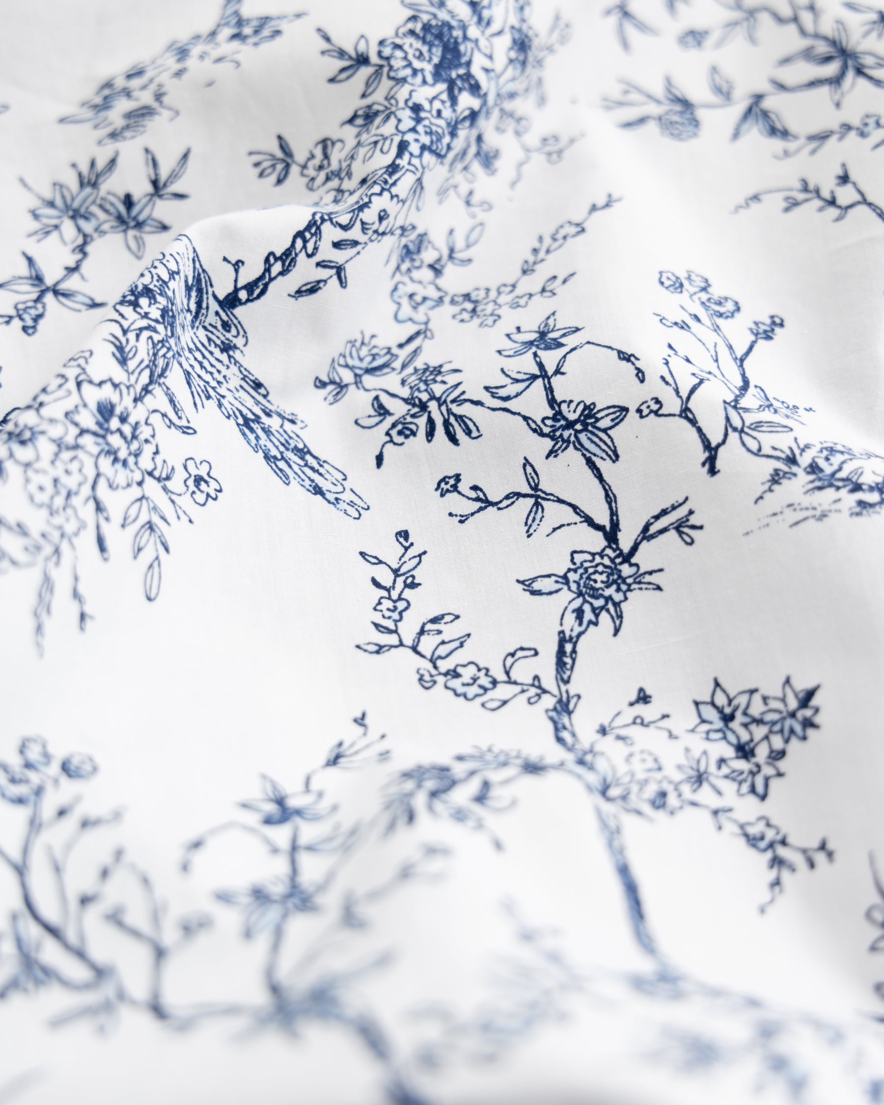 The Luxe Premium Cotton Bed Sheets in Timeless Toile by Petite Plume feature a close-up of white fabric adorned with an elegant blue floral and branch pattern. This delicate design, reminiscent of luxury bedding, exudes botanical elegance with intricate blossoms and leaves for a calming effect.
