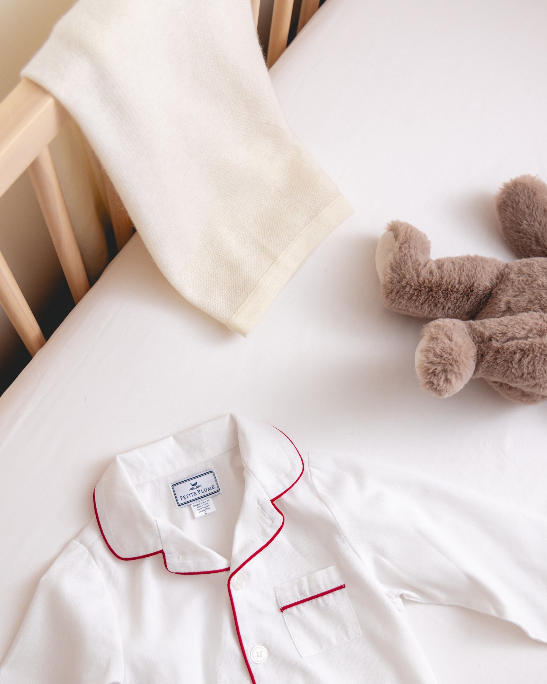 A childrens crib features a Signature Luxe Sateen Crib Sheet in white by Petite Plume, a soft off-white blanket draped over the rail, a plush gray stuffed animal near the pillow, and white pajamas with red piping neatly laid out.