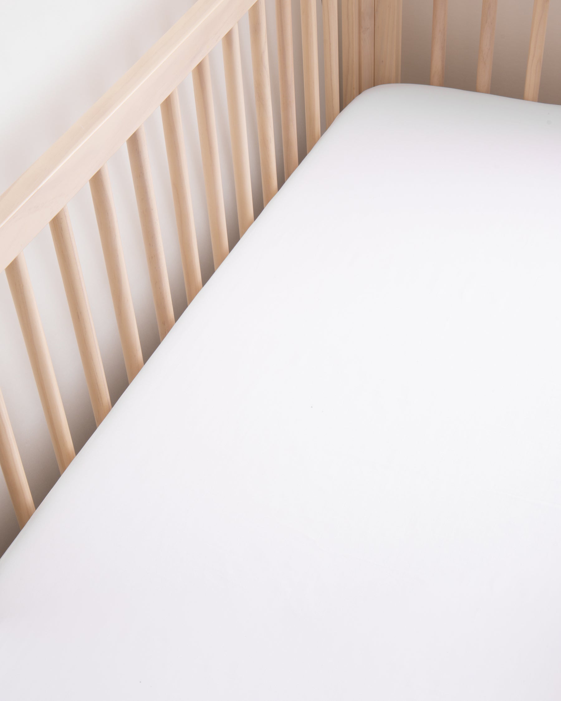 A wooden crib with a natural finish features the Signature Luxe Sateen Crib Sheet in White by Petite Plume on the mattress. It has vertical slats on the sides, and an above view highlights its smooth, clean surface.