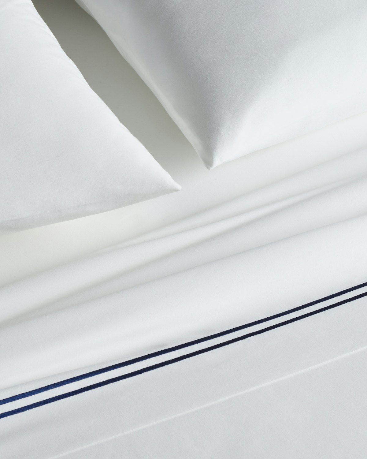 Close-up of a neatly made bed with Petite Plume Luxe Premium Cotton Sheets in White with Navy. The crisp percale sheets boast two thin, parallel dark blue stripes near the edge, crafted from long-staple cotton. Matching pillowcases complete the elegant set.