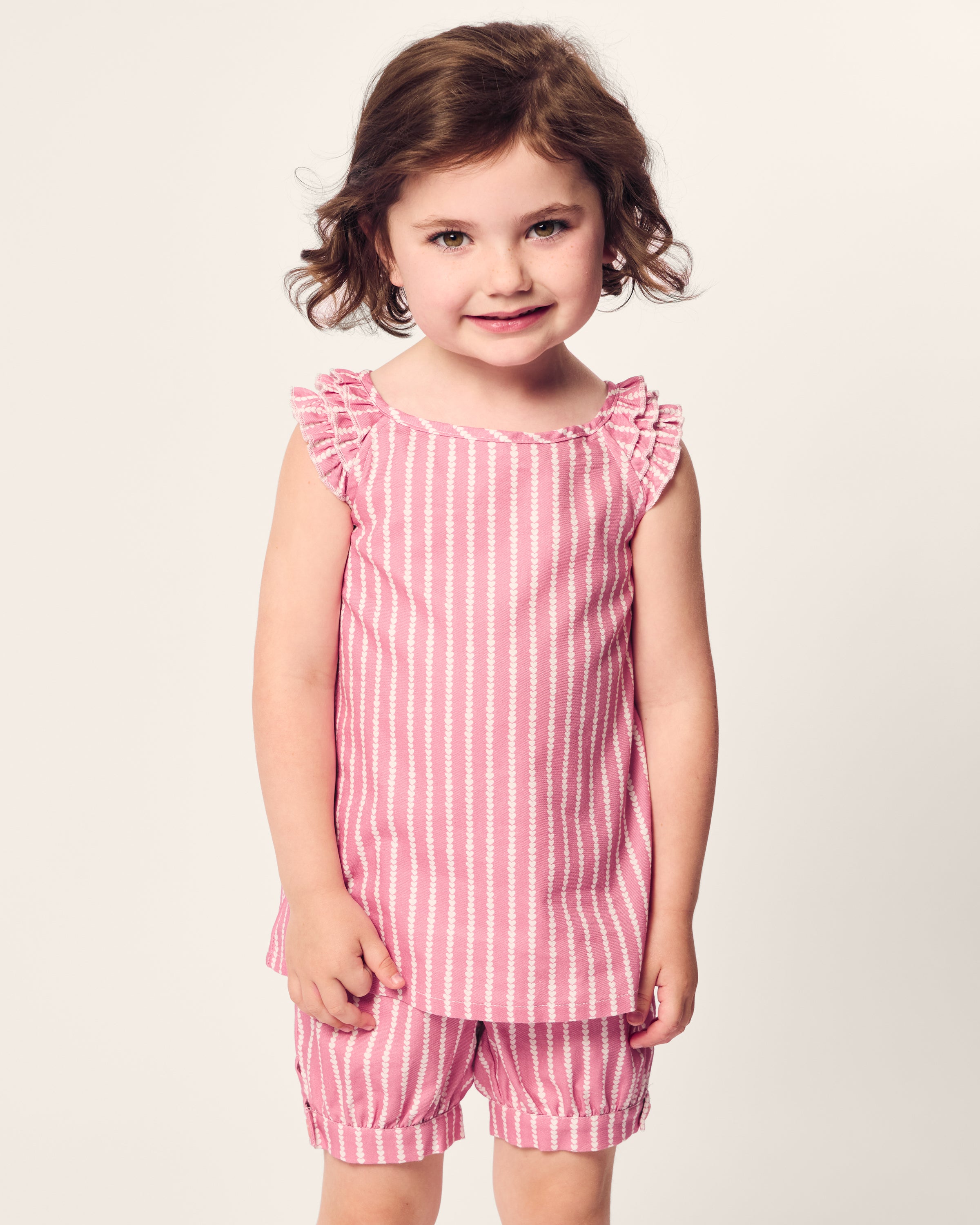 A young child with curly brown hair is smiling, wearing Petite Plumes Girls Twill Amelie Short Set in Love Lines, featuring a pink and white striped top with ruffled sleeves and matching shorts. The background is a plain light color.