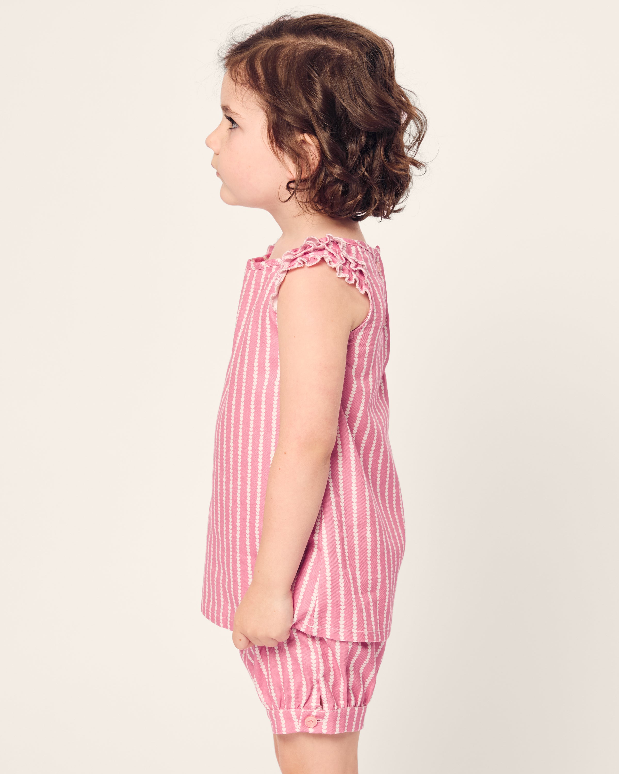 A young child with curly brown hair stands in profile, facing left, wearing the Girls Twill Amelie Short Set in Love Lines by Petite Plume. The set features a pink and white striped sleeveless top with ruffled edges and matching cotton shorts. The background is plain and neutral.