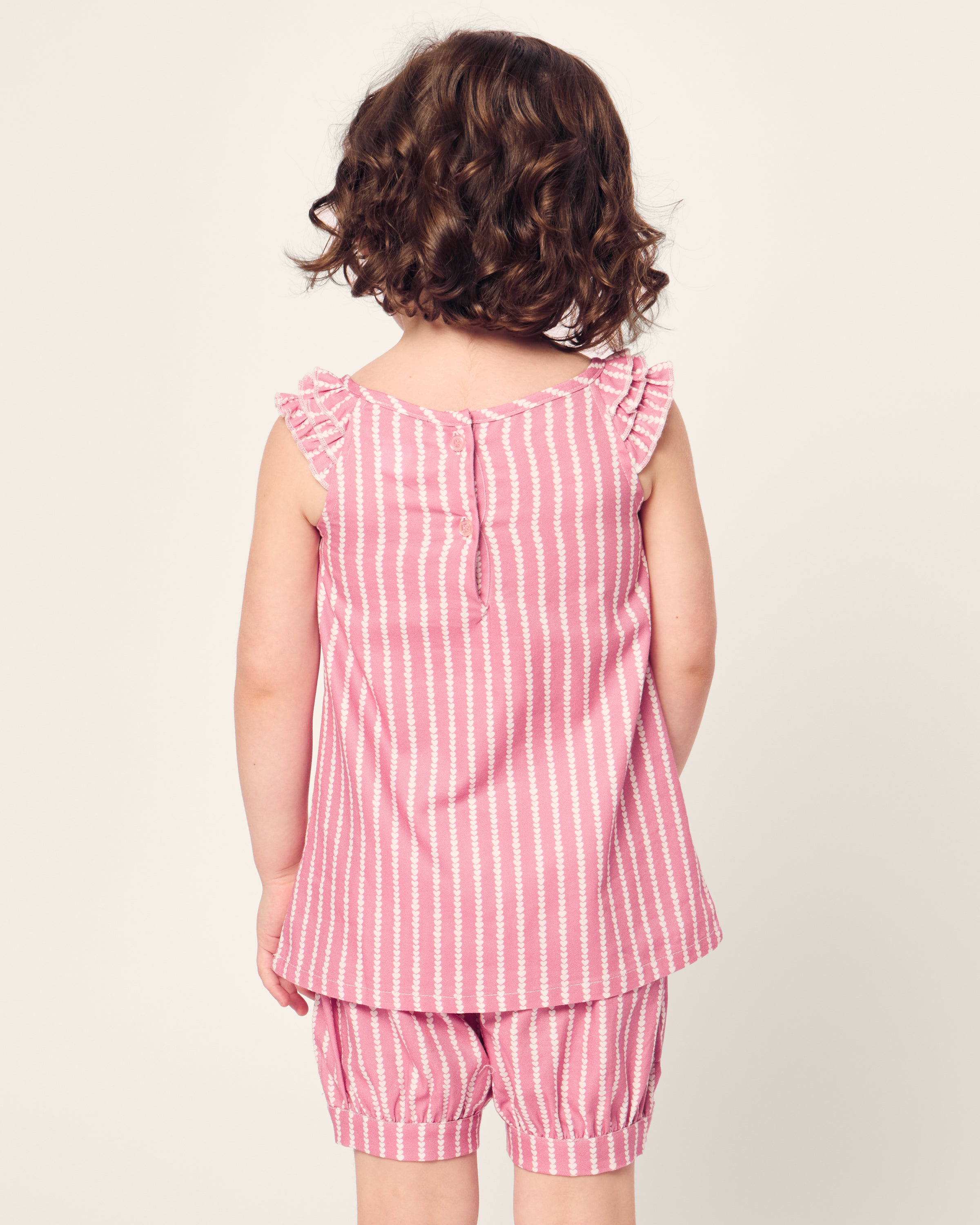 A young child with curly hair faces away, wearing Petite Plumes Girls Twill Amelie Short Set in Love Lines. The pink and white striped sleeveless top features ruffled shoulders and matching shorts, crafted from soft cotton sleepwear material, set against an off-white background.