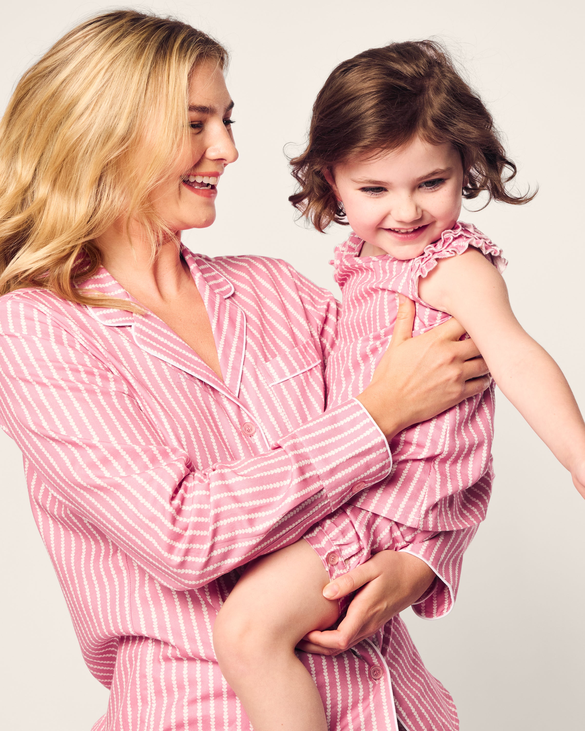 A smiling woman with long blonde hair wears luxurious pink and white striped pajamas, holding a cheerful child dressed in Petite Plumes Girls Twill Amelie Short Set in Love Lines. Both stand against a plain background.