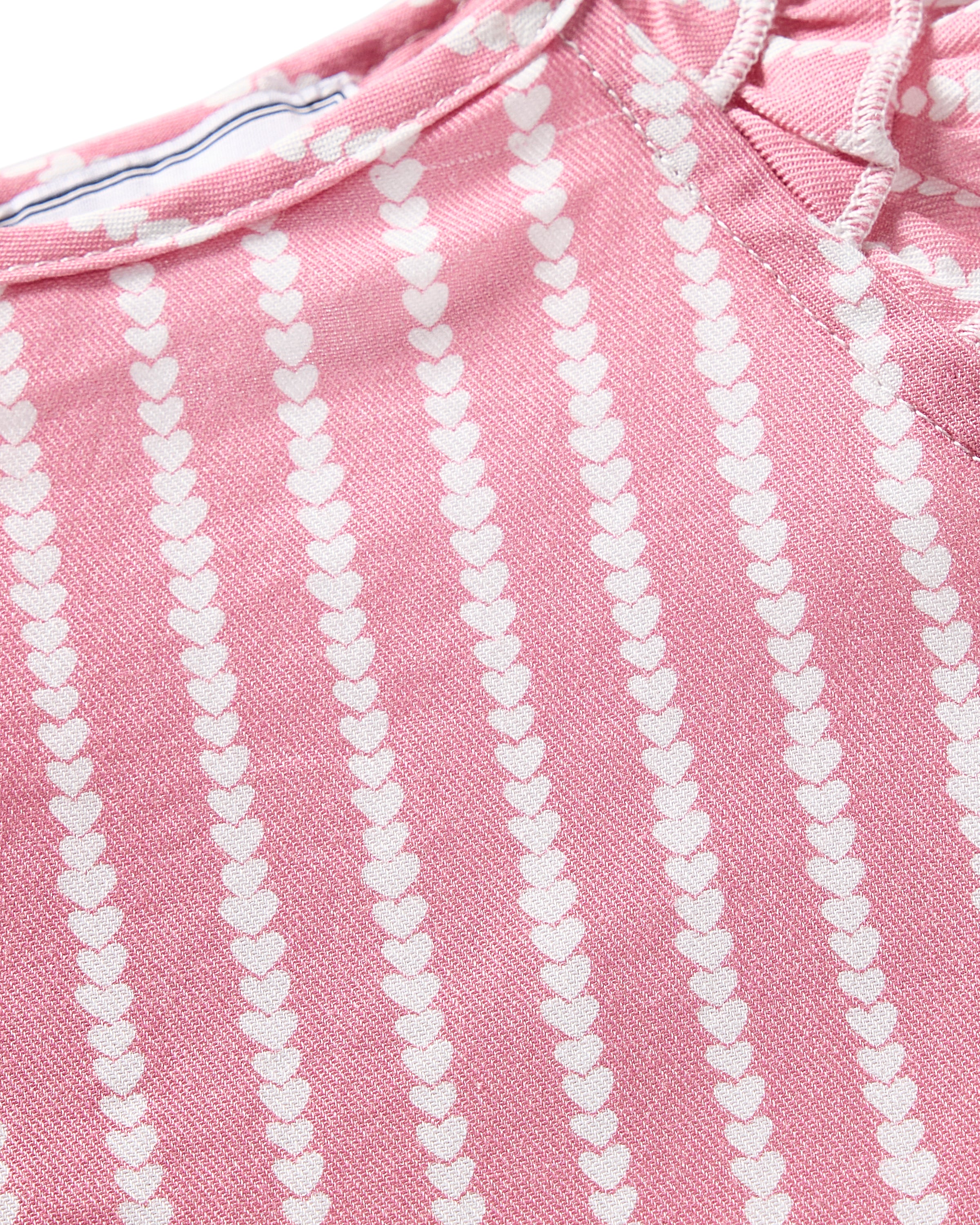 A close-up of pink fabric with vertical rows of small white hearts suggests it is part of the luxurious Girls Twill Amelie Short Set in Love Lines by Petite Plume, featuring a slightly textured material and visible seams indicative of premium sleepwear.