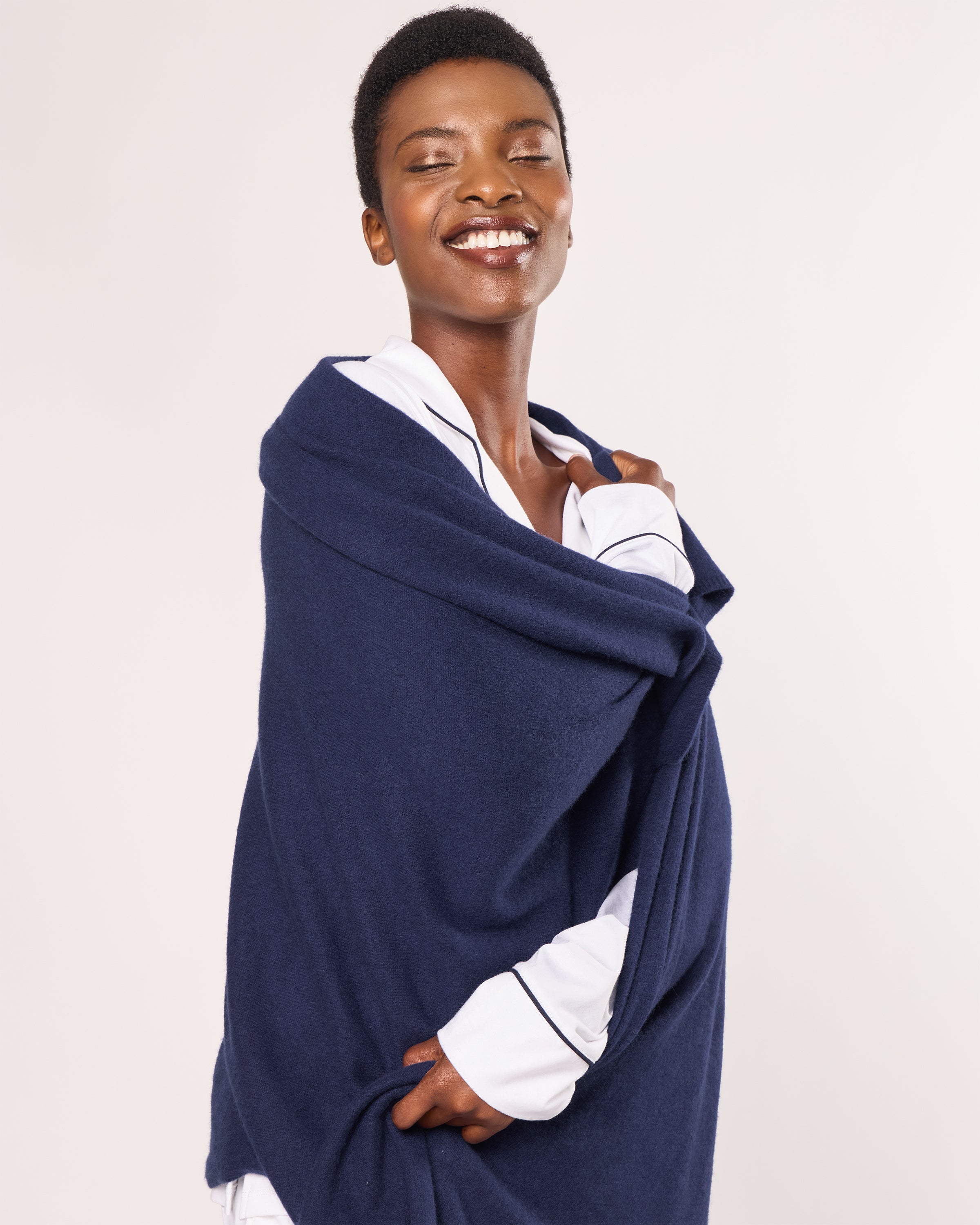 A person smiles warmly, draped in the luxurious Petite Plume Womens Cashmere Wrap in Navy over a white shirt. The plain, light background accentuates their joyful expression and cozy, casual vibe.