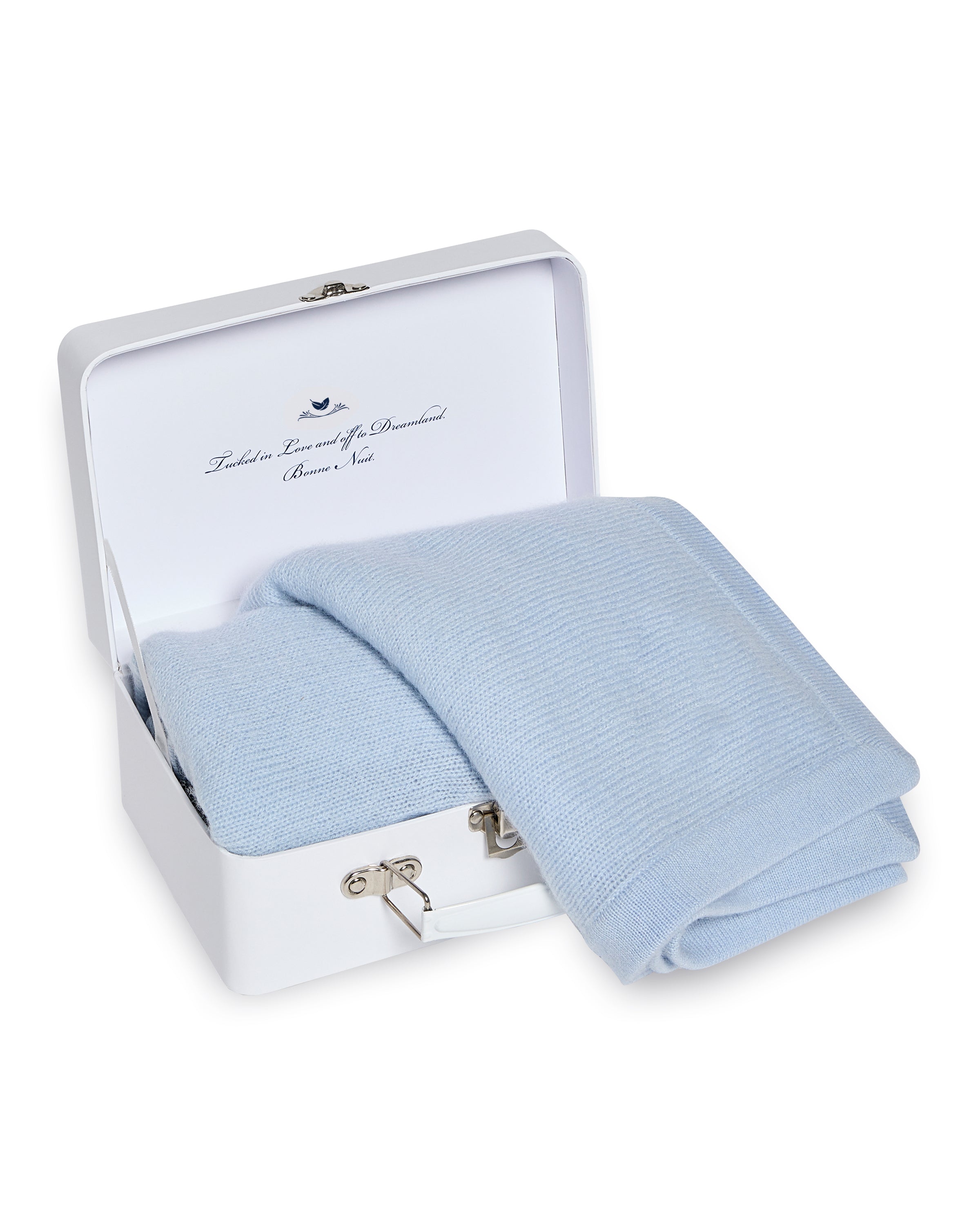 A Petite Plume Indigo cashmere baby blanket is neatly folded inside a white suitcase, which is open to reveal text on the lid and has a metallic latch in front. Handmade in Nepal, this elegant piece makes a perfect luxury gift.