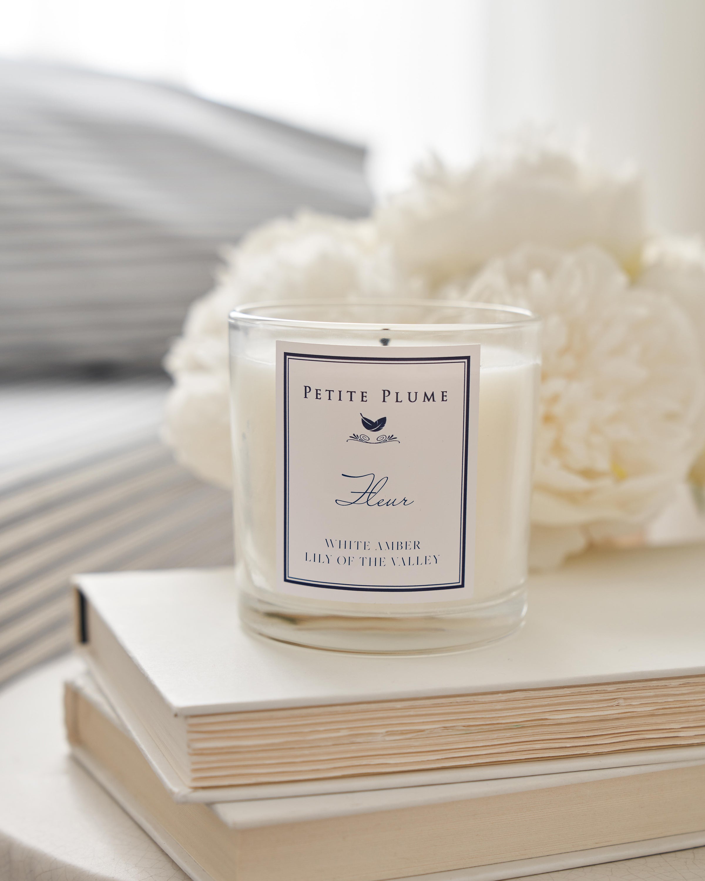 A white Petite Plume Fleur candle from the Petite Plume Collection sits on books, exuding a White Amber & Lily of the Valley scent. A softly focused white flower glows in gentle light behind it.