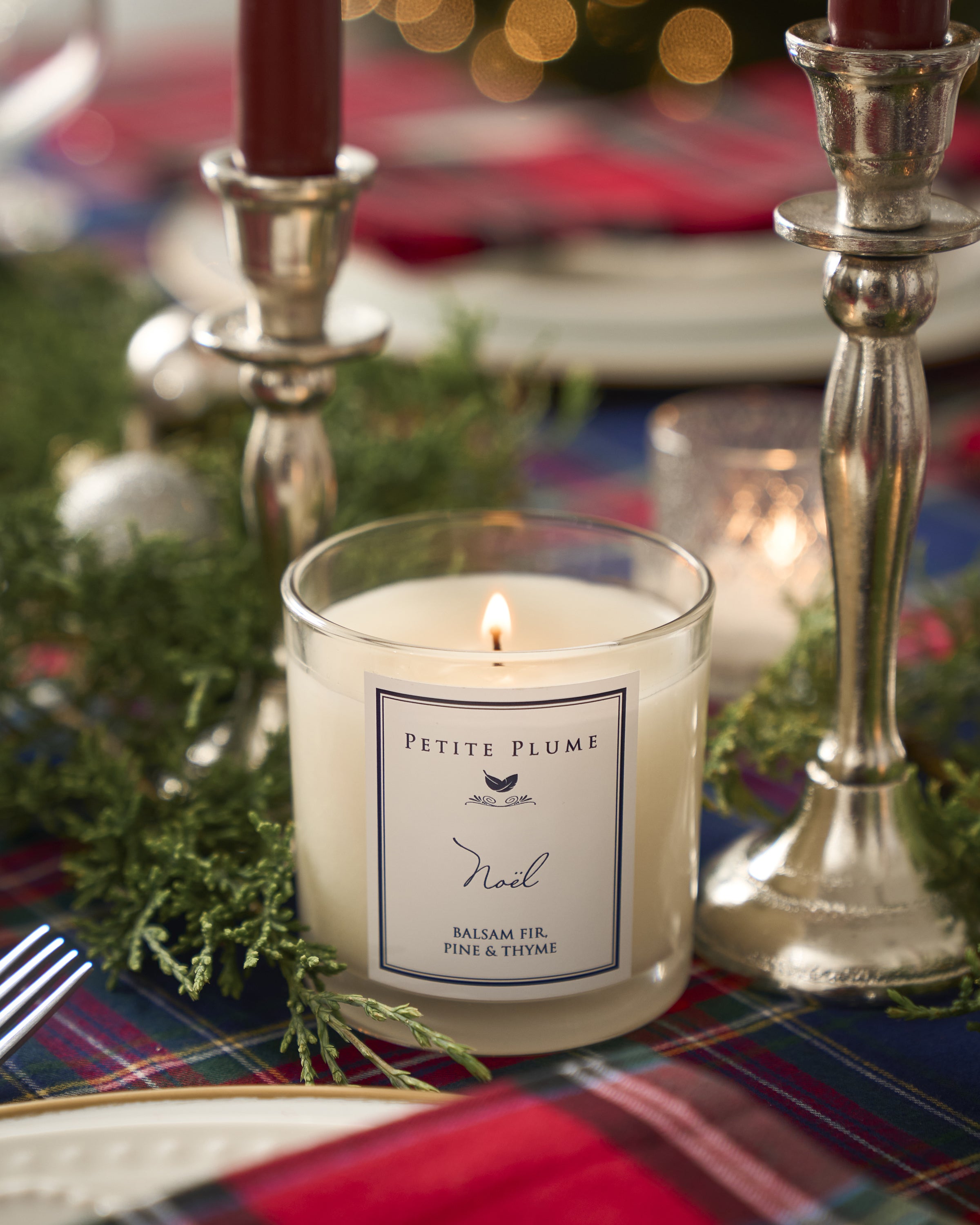 The Petite Plume Noël Candle from the Petite Plume brand, with its Balsam Fir, Pine & Thyme scent, enhances a festive table adorned with plaid cloth, silver candlesticks, greenery, and red candles, creating a cozy holiday atmosphere.