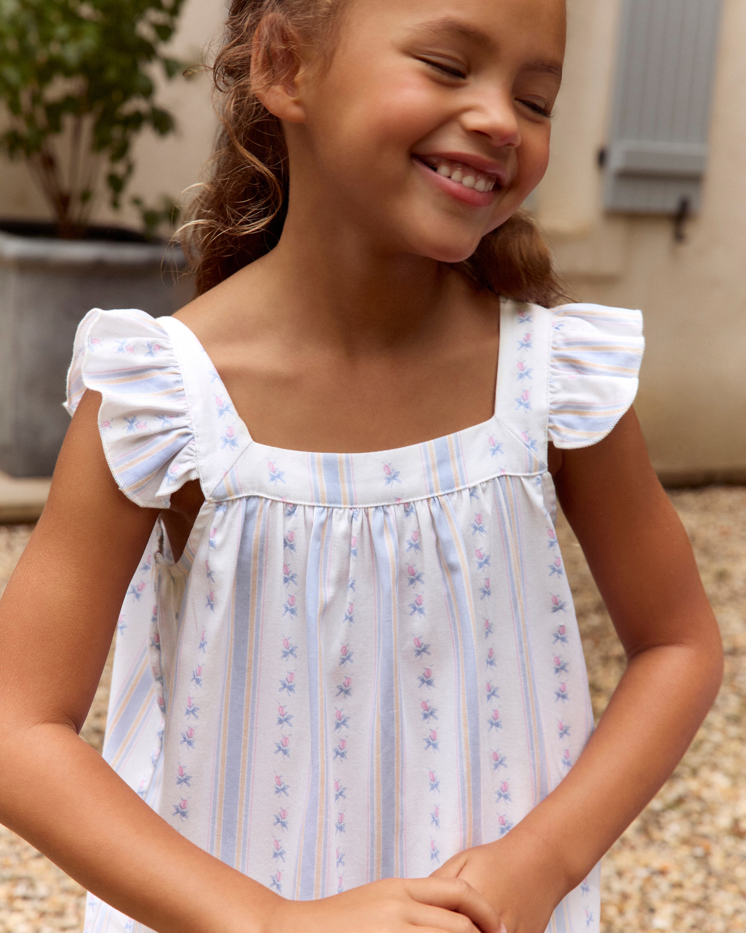 A young girl with long dark hair stands barefoot, smiling, wearing the Girl's Twill Clara Nightgown in Cottage Stripe—white with subtle pastel stripes and ruffled shoulders—against a plain light background.